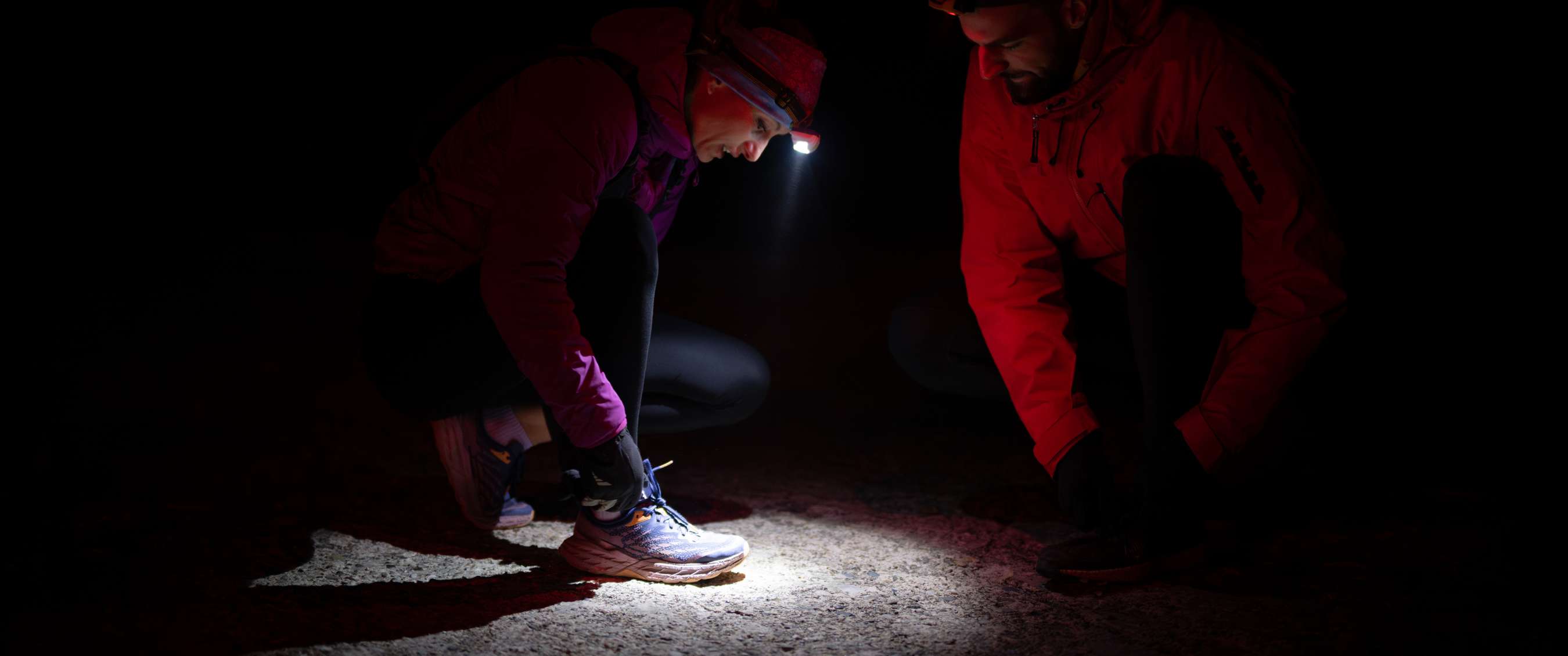 Deux coureurs sont accroupis dans l'obscurité tandis que l'un d'eux noue ses lacets, éclairés par une lampe frontale.