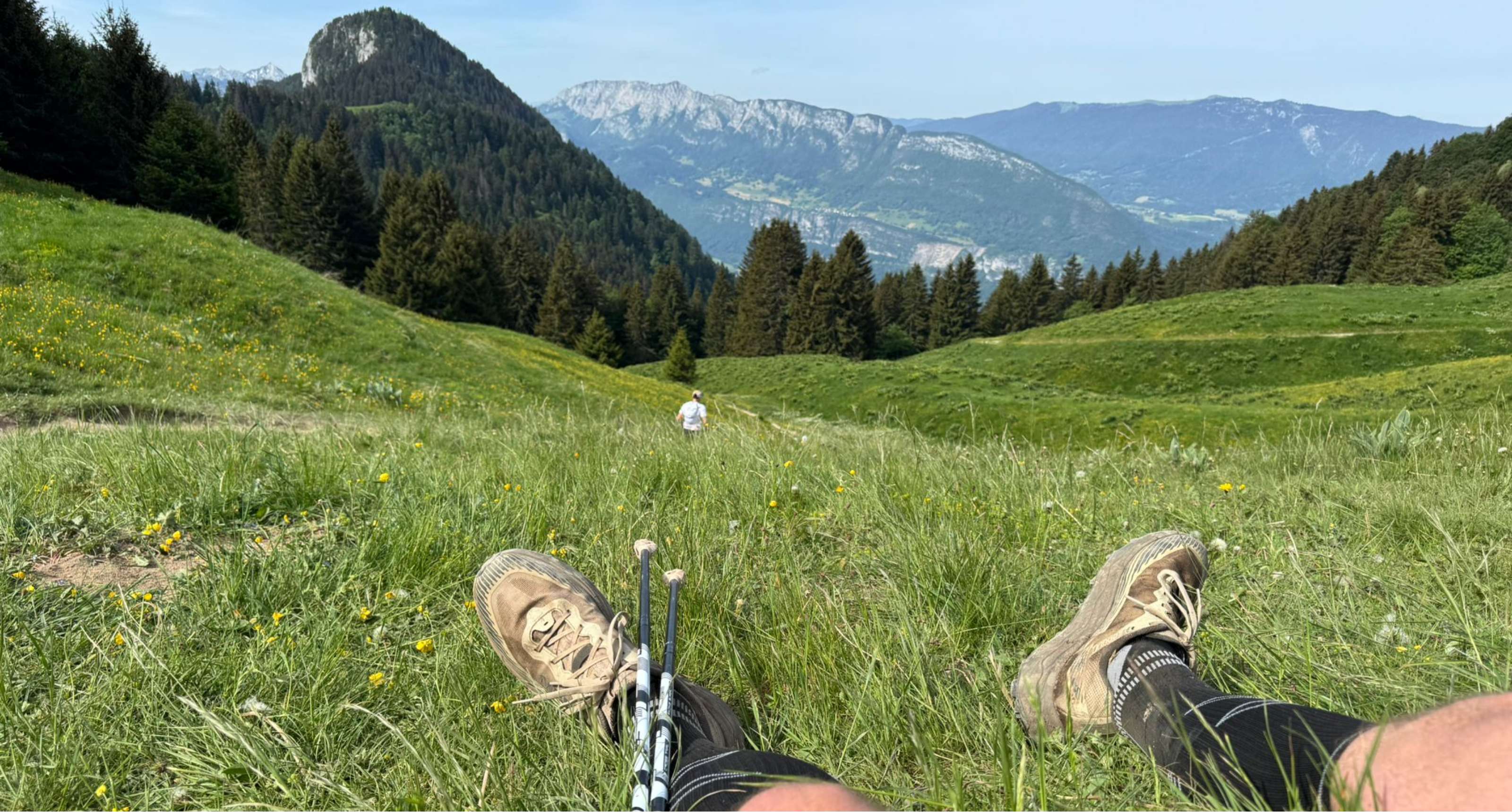Une personne se repose dans l’herbe avec ses chaussures et bâtons face au paysage montagneux.