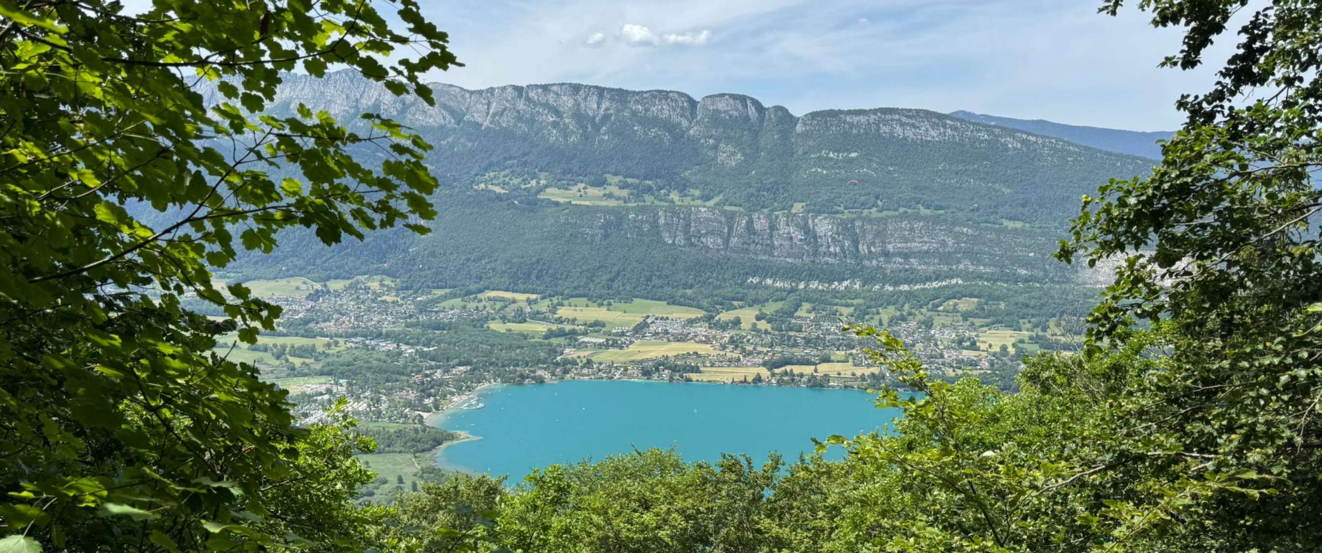 Vue panoramique sur un lac turquoise, une vallée et des montagnes à travers des branches vertes.