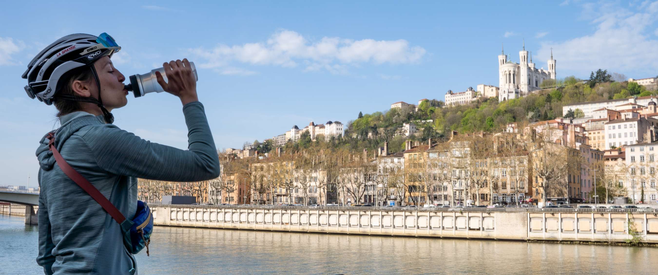 Persoon drinkt uit een bidon aan het water, met stad en basiliek op de achtergrond.