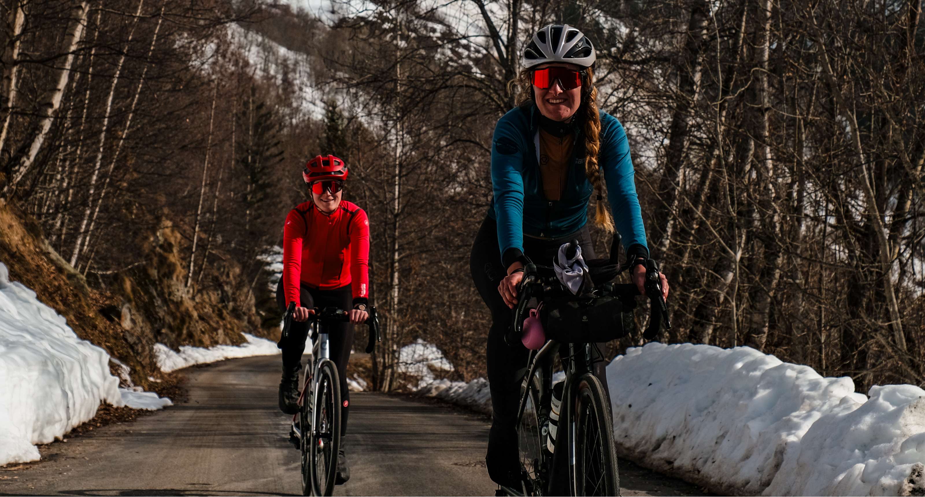Une cycliste en bleu précède une cycliste en rouge sur une route forestière bordée de neige.