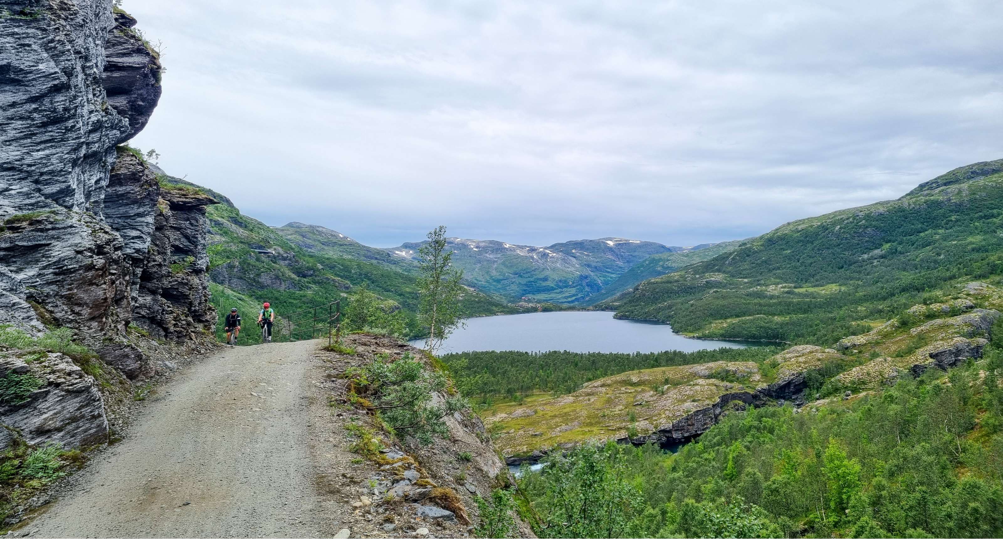 Deux cyclistes roulent sur un chemin de gravier le long d'une paroi rocheuse, face à un lac de montagne.