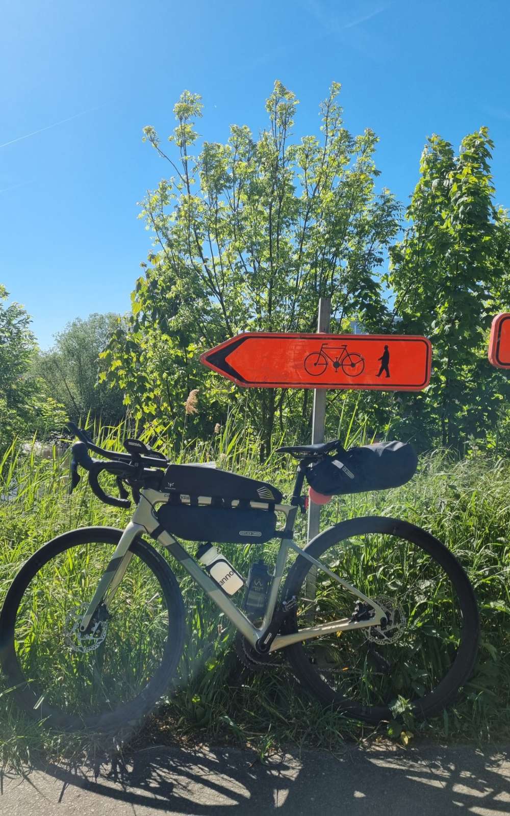 Een fiets met frame- en zadeltassen staat in het gras onder een rode wegwijzer voor fietsers en wandelaars.
