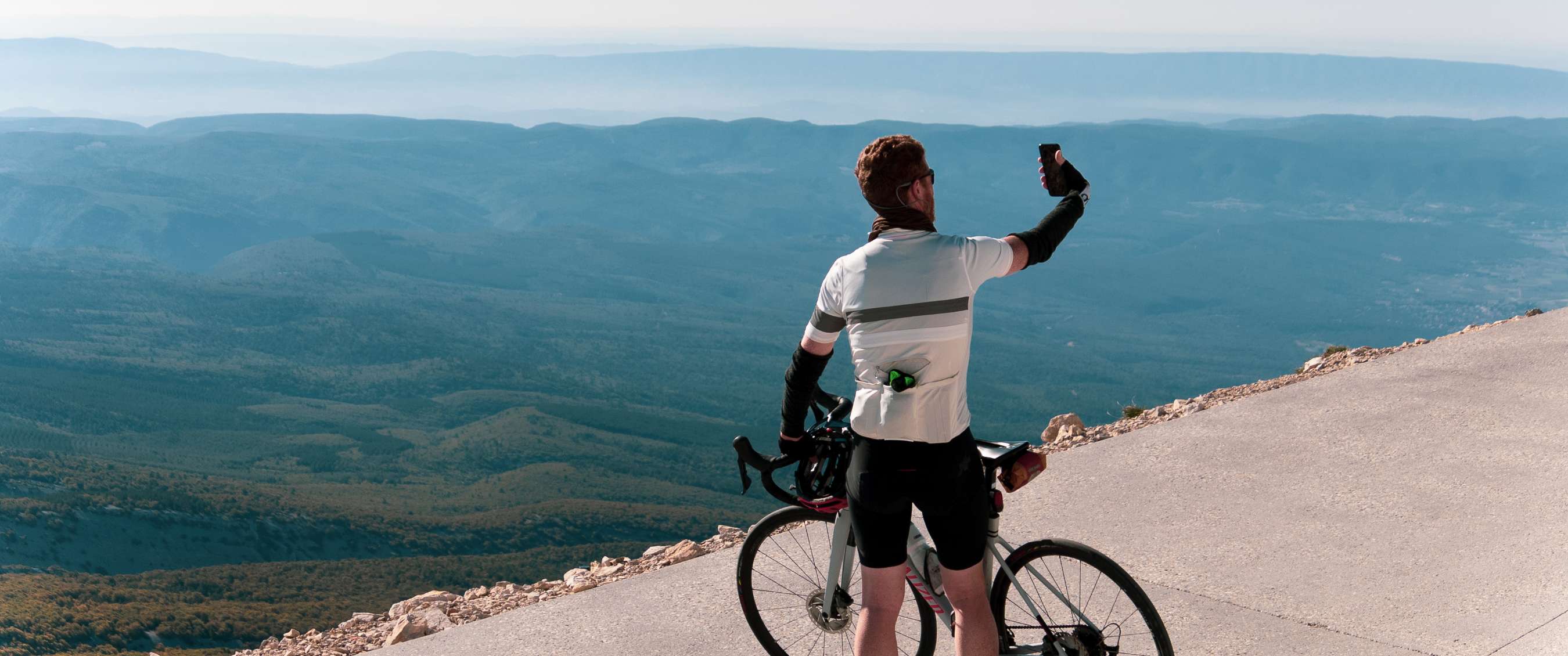 Cycliste prenant un selfie à côté de son vélo sur une route de montagne avec vue panoramique.