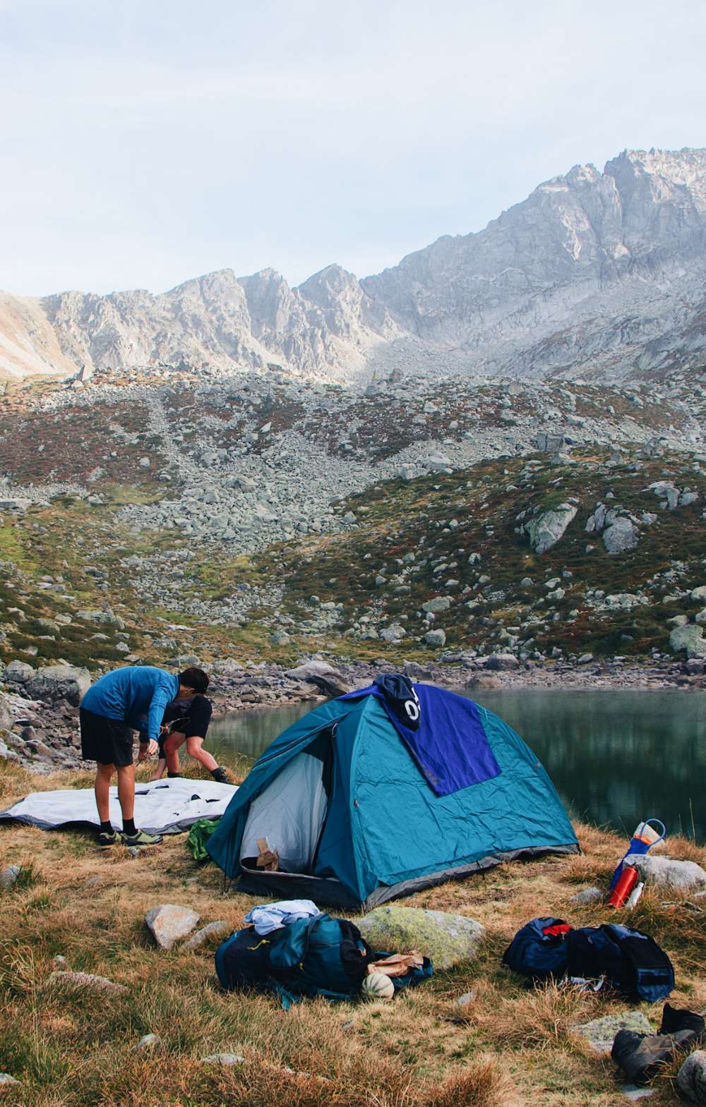 Tente bleue au bord d’un lac de montagne, avec deux personnes déballant du matériel