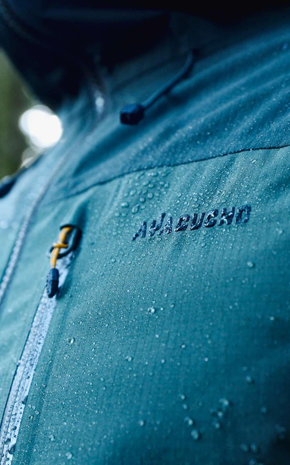 Waterdruppels liggen op een blauwe outdoorjas met rits en zichtbaar Ayacuchos-logo.