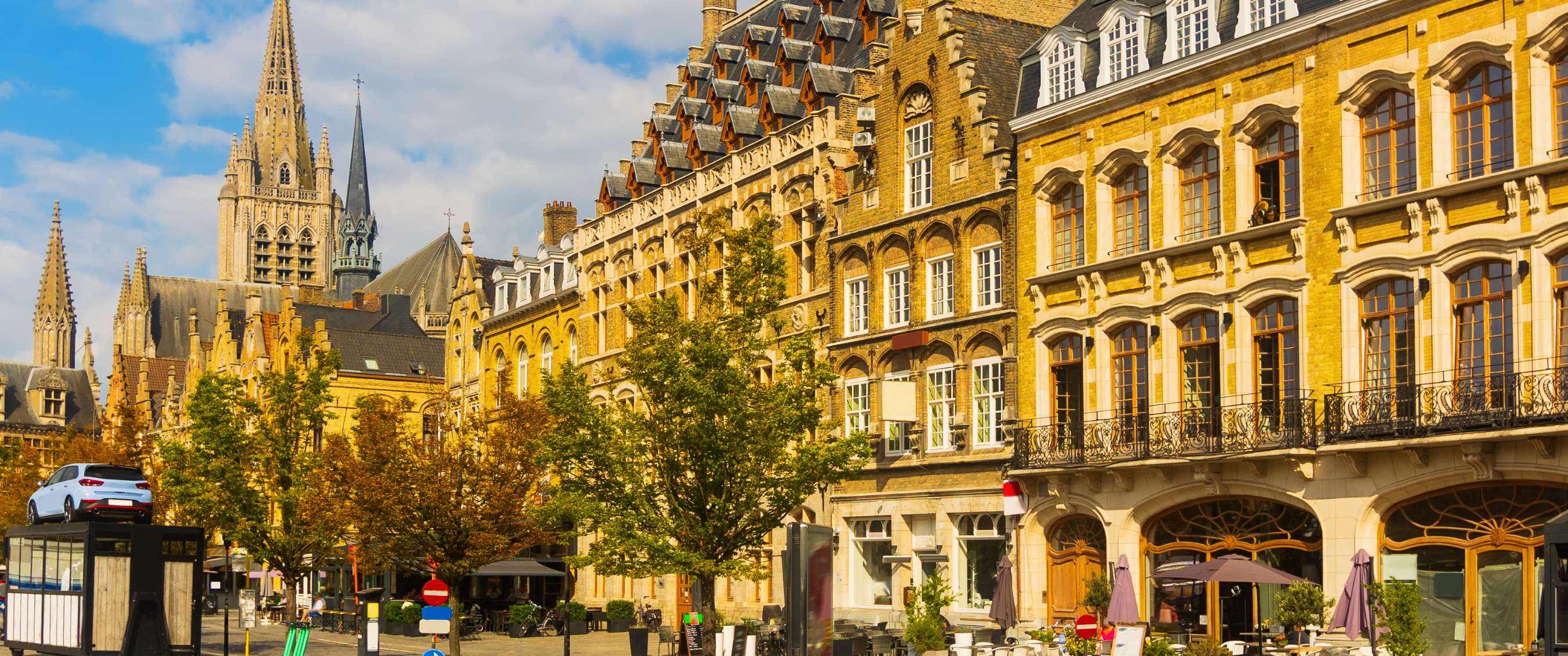 Rue historique avec haute tour d’église, façades ornées et arbres sous un ciel nuageux.