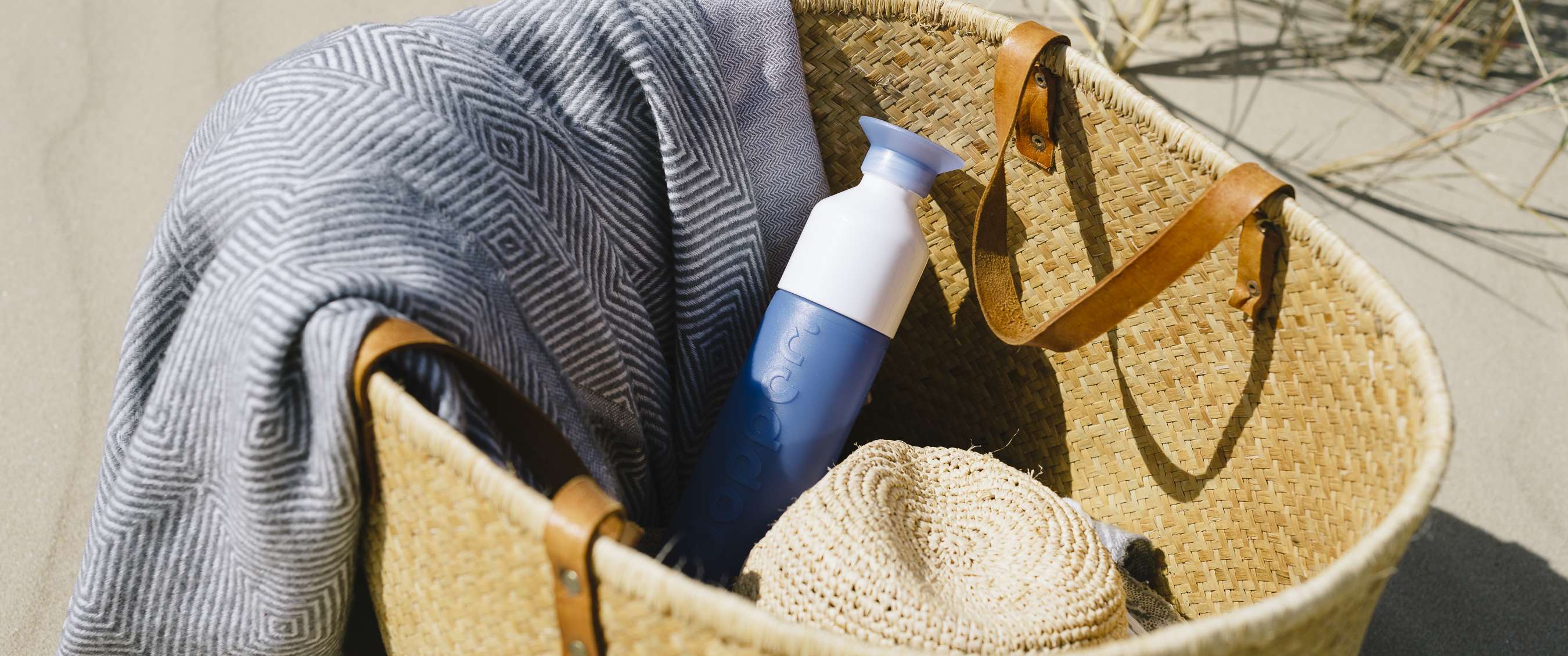 Blauwe Dopper-drinkfles ligt in een gevlochten strandtas met handdoek en hoed.