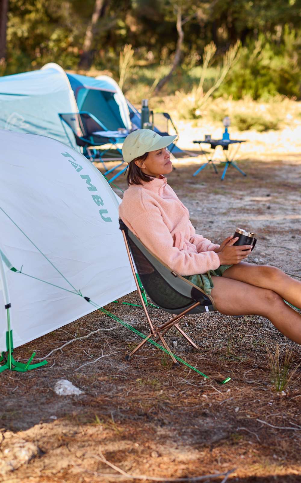 Personne assise avec une tasse dans une chaise de camping près d’une tente blanche TAMBU.