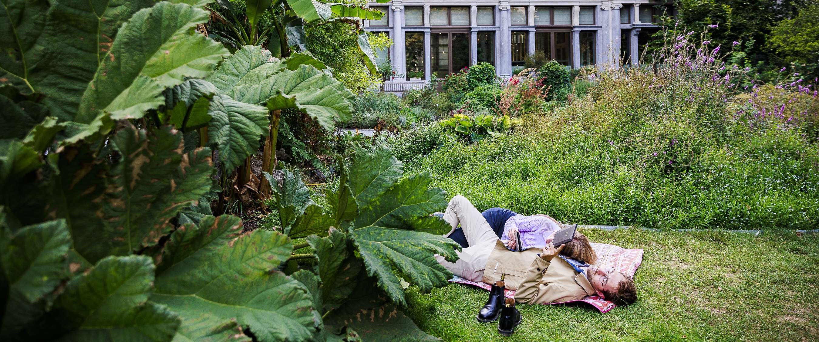 Deux personnes allongées sur la pelouse dans un parc urbain verdoyant avec bâtiment historique en arrière-plan.