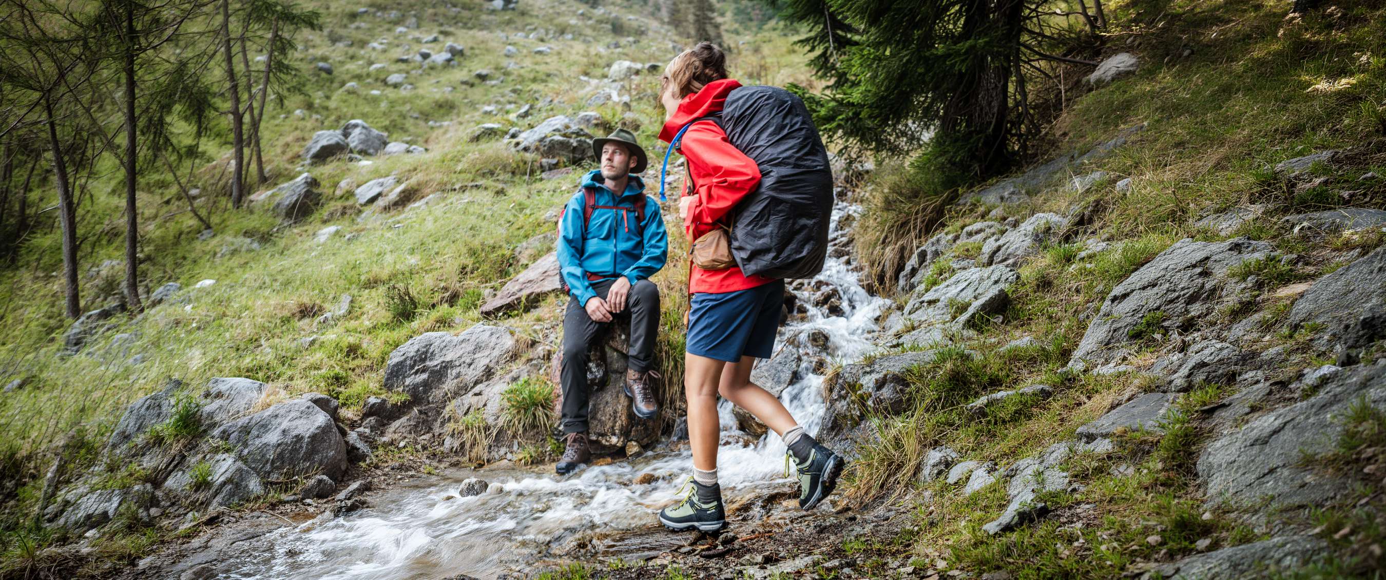 Wandelaars met hoge wandelschoenen bij stromend water tussen rotsen en naaldbomen.