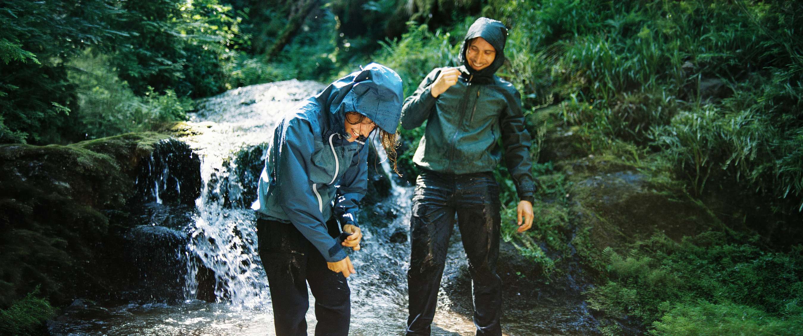 Twee personen in waterdichte kleding staan bij een waterval tussen groene begroeiing.