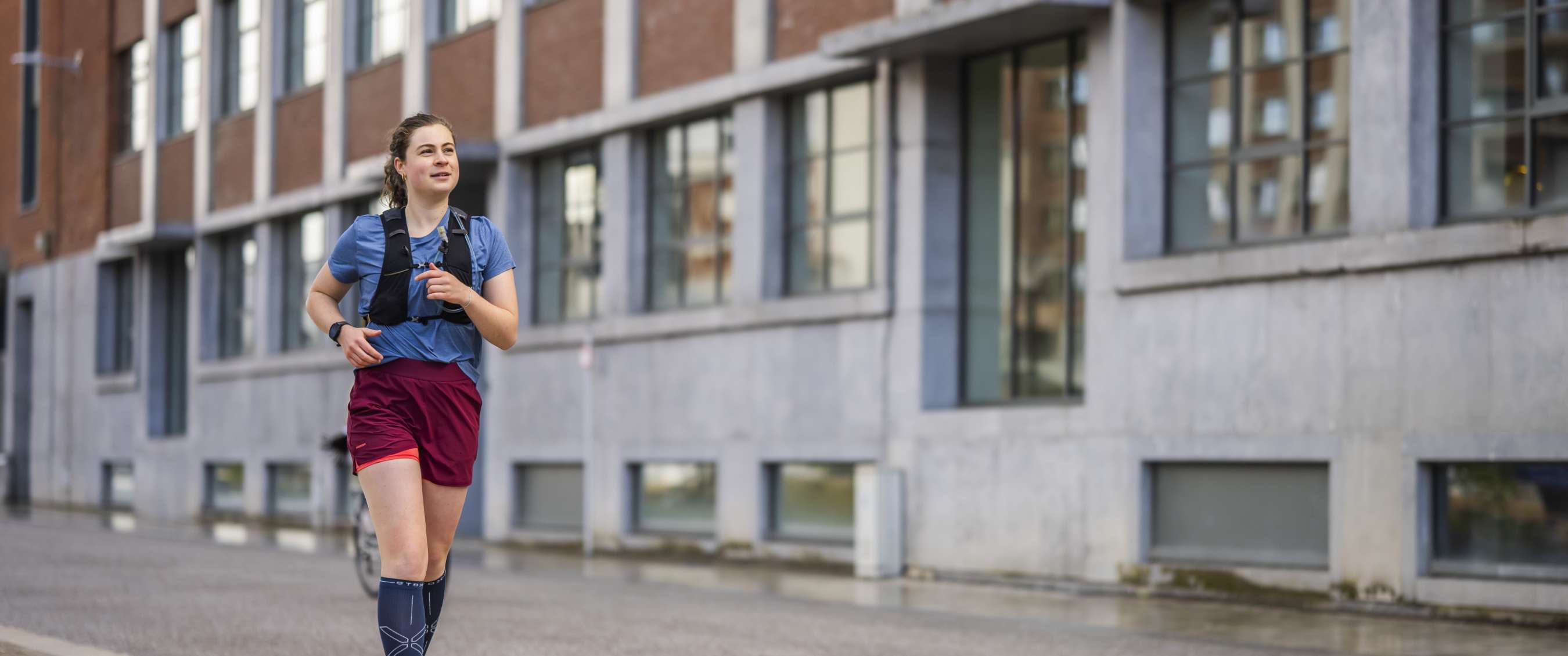 Vrouw loopt hard in de stad langs een gebouw, met blauw shirt, bordeaux short en zwarte loopvest.