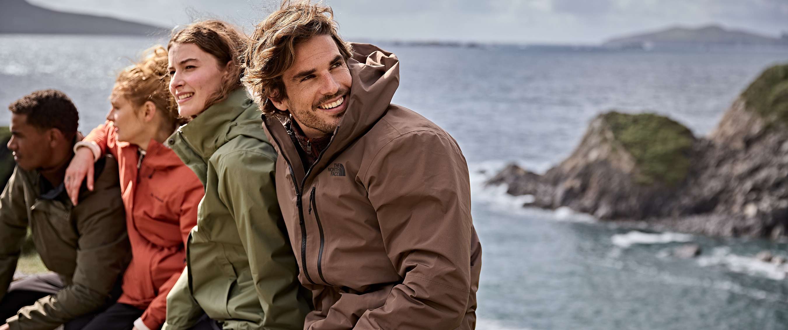Deux femmes et deux hommes en vestes colorées se détendent au bord de la mer avec collines en arrière-plan.