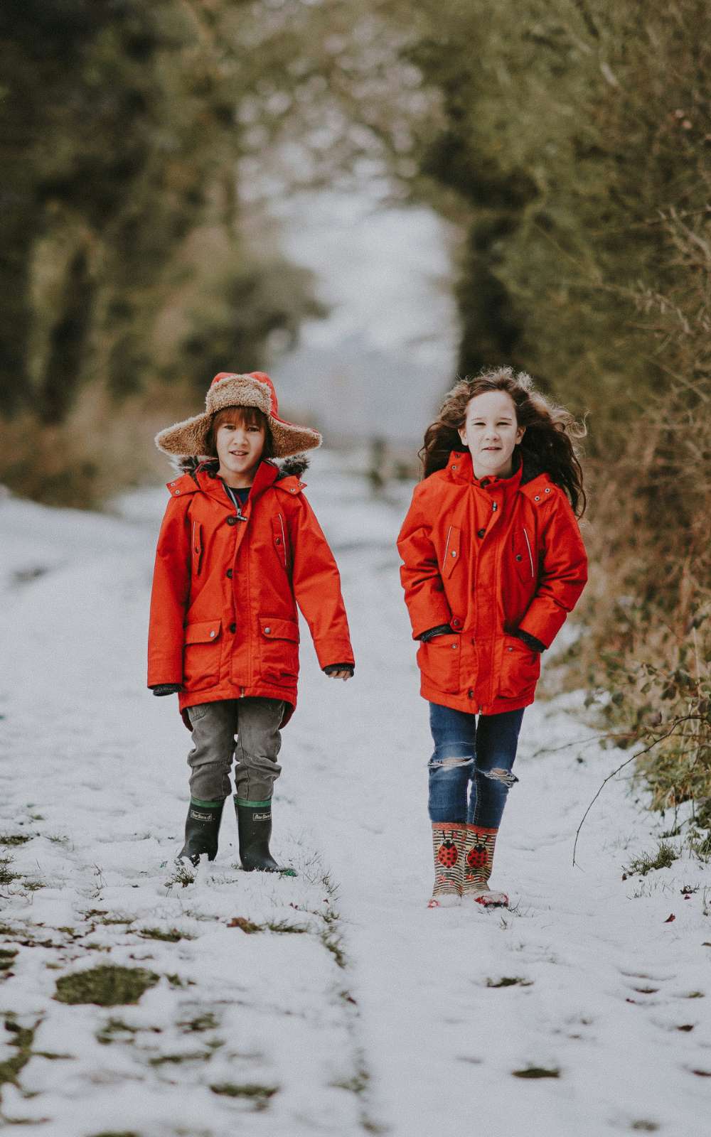 Twee kinderen in rode winterjassen en regenlaarzen wandelen over een besneeuwd bospad.