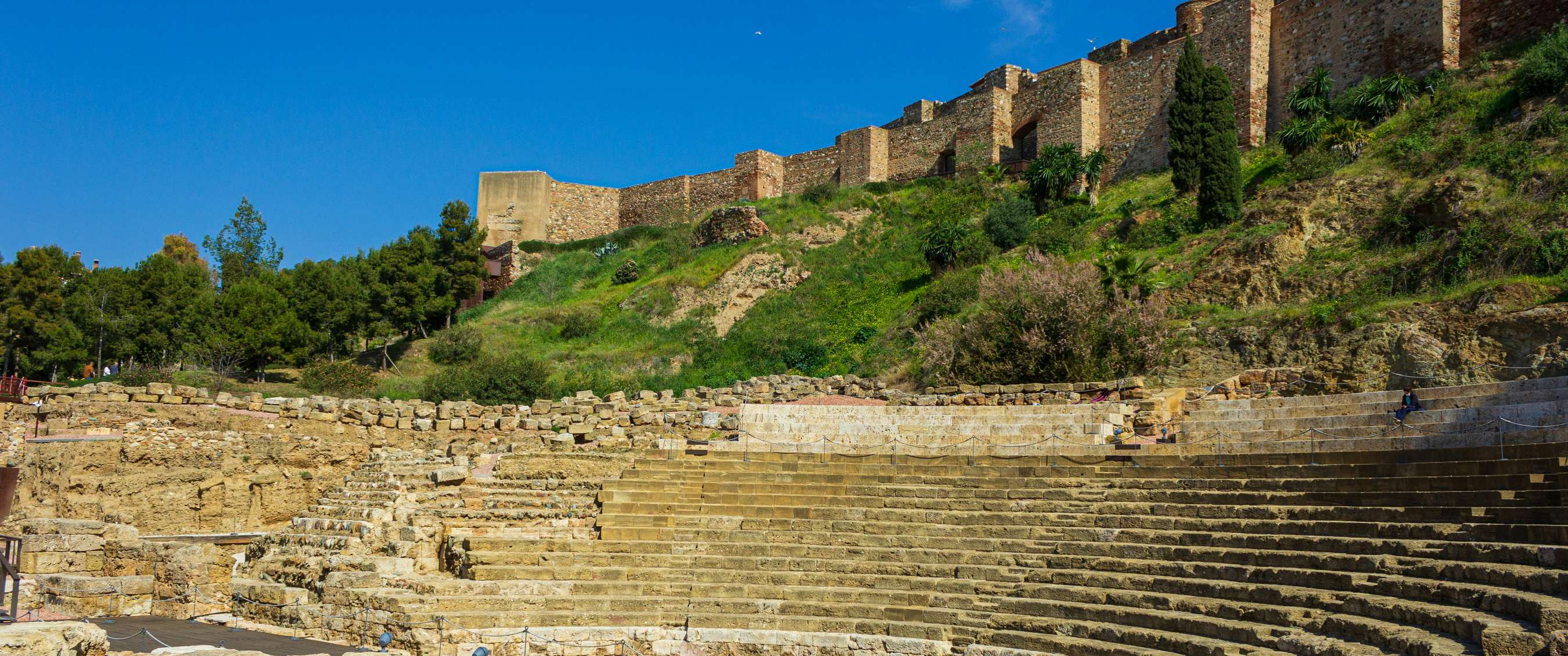 Roman theatre with stone seating and fortress walls on a green hillside in Málaga.