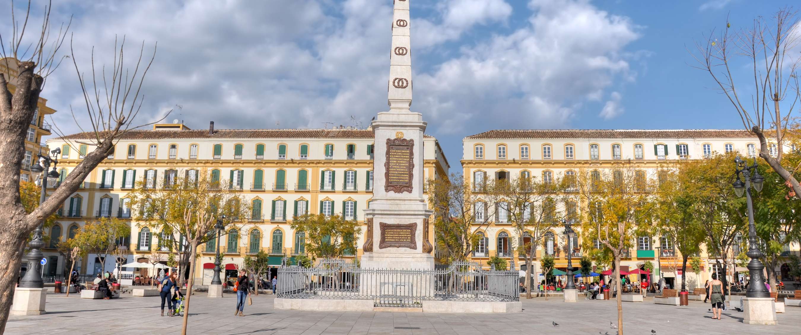 White obelisk on a spacious square in Málaga, surrounded by historic façades and trees.