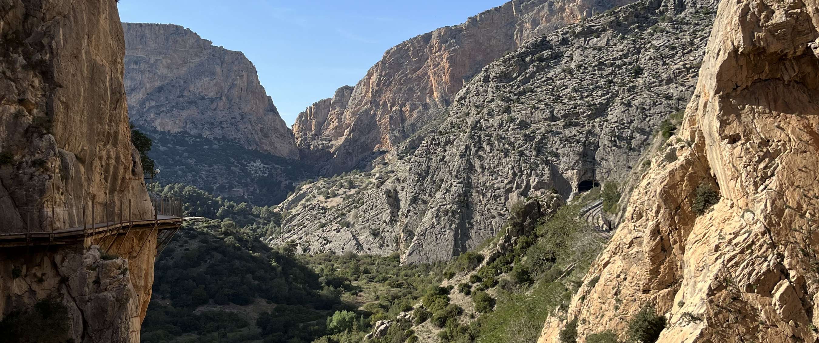 Narrow walkway along steep rock walls in a green mountain gorge beneath a clear blue sky.