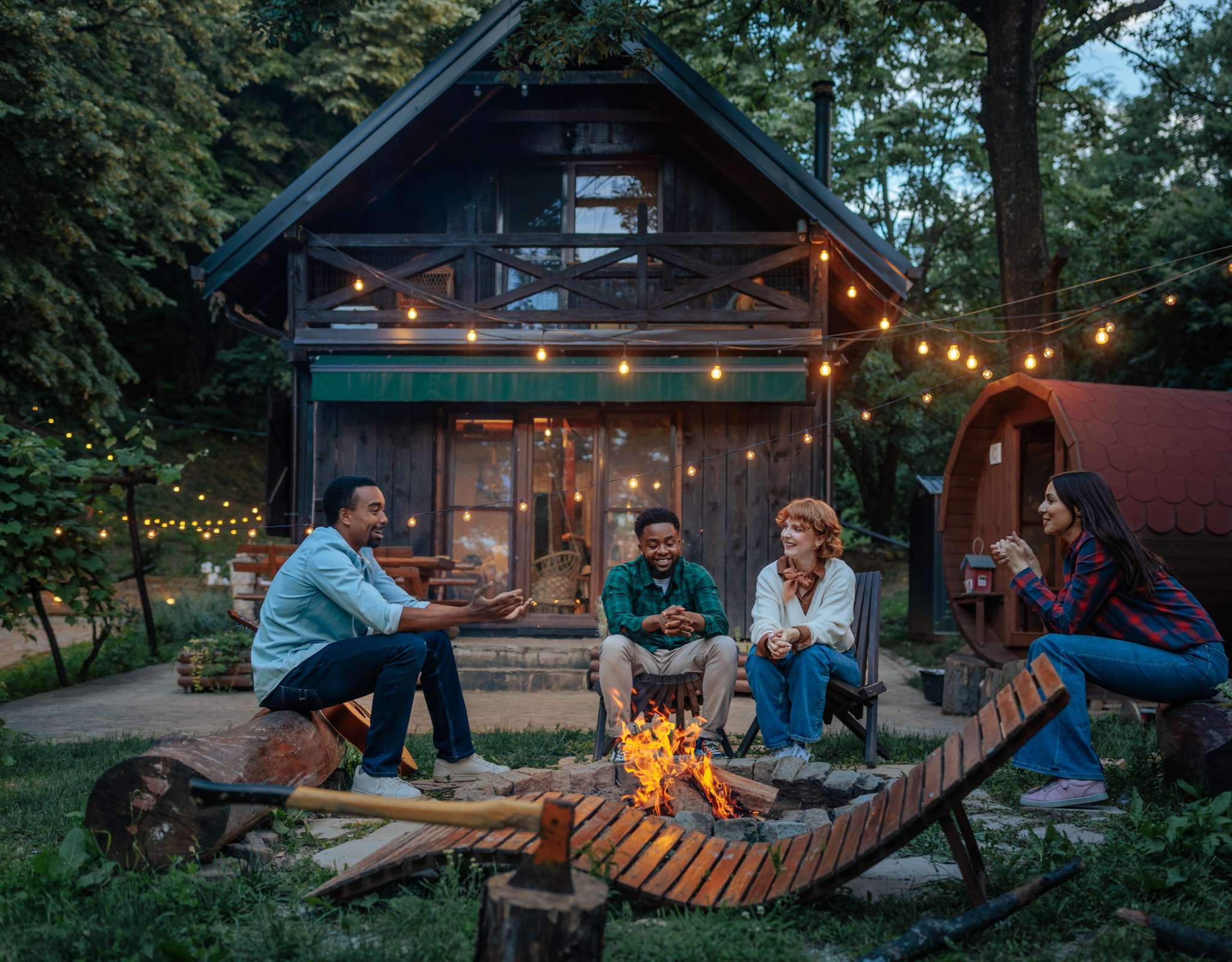 Vier mensen zitten bij een kampvuur voor een houten hut met hangende lichtjes.