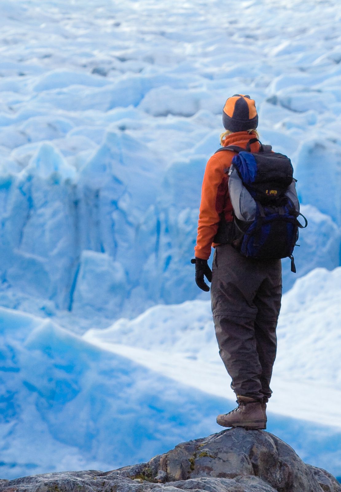 Wandelaar met oranje jas kijkt uit over een uitgestrekte gletsjer.