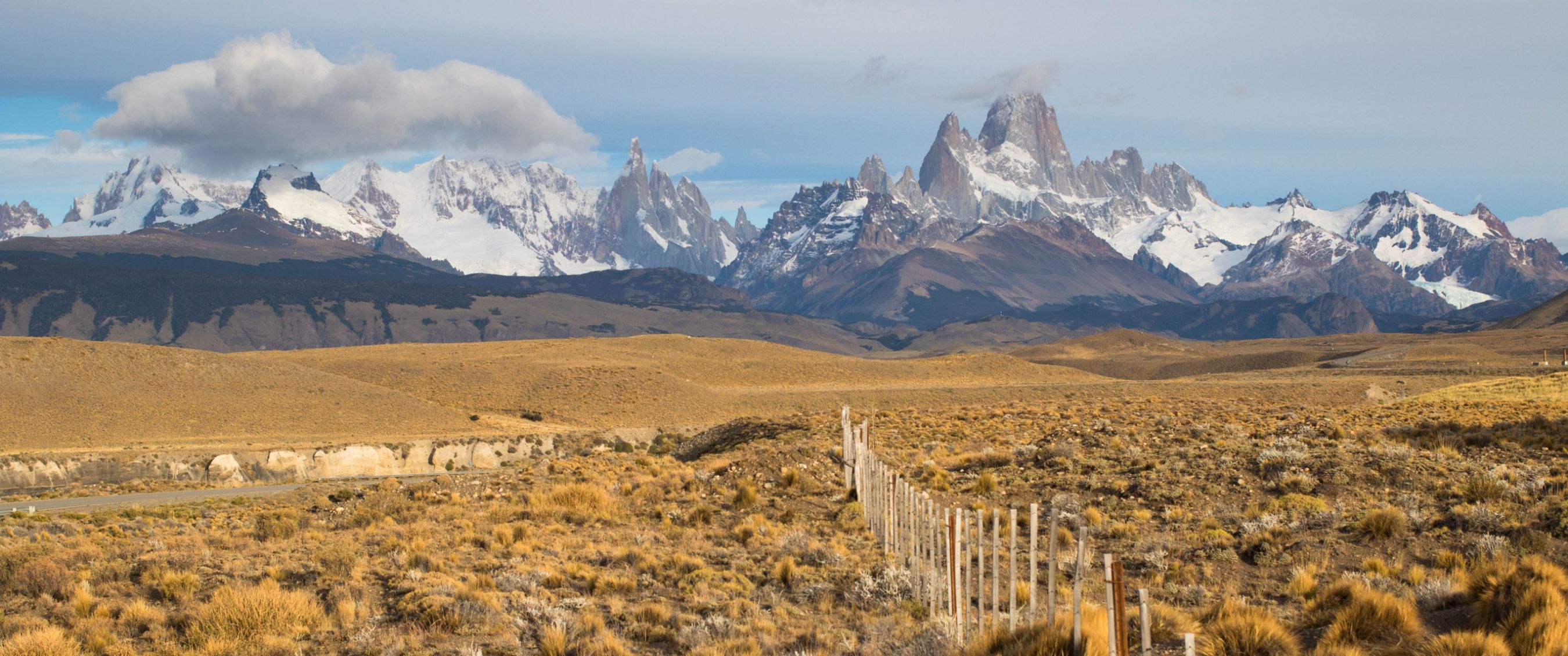 Uitgestrekt steppegebied met houten hek en besneeuwde bergtoppen aan de horizon.