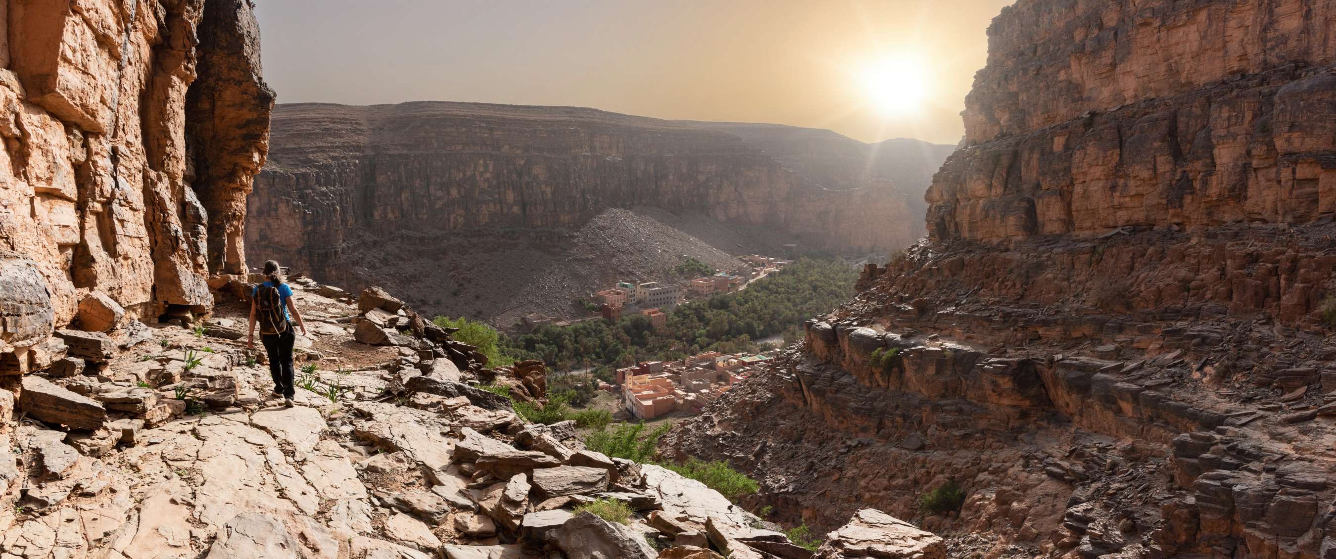 Wandelaar met rugzak op smal rotsachtig pad in een kloof met zicht op bergdorp bij laagstaande zon.
