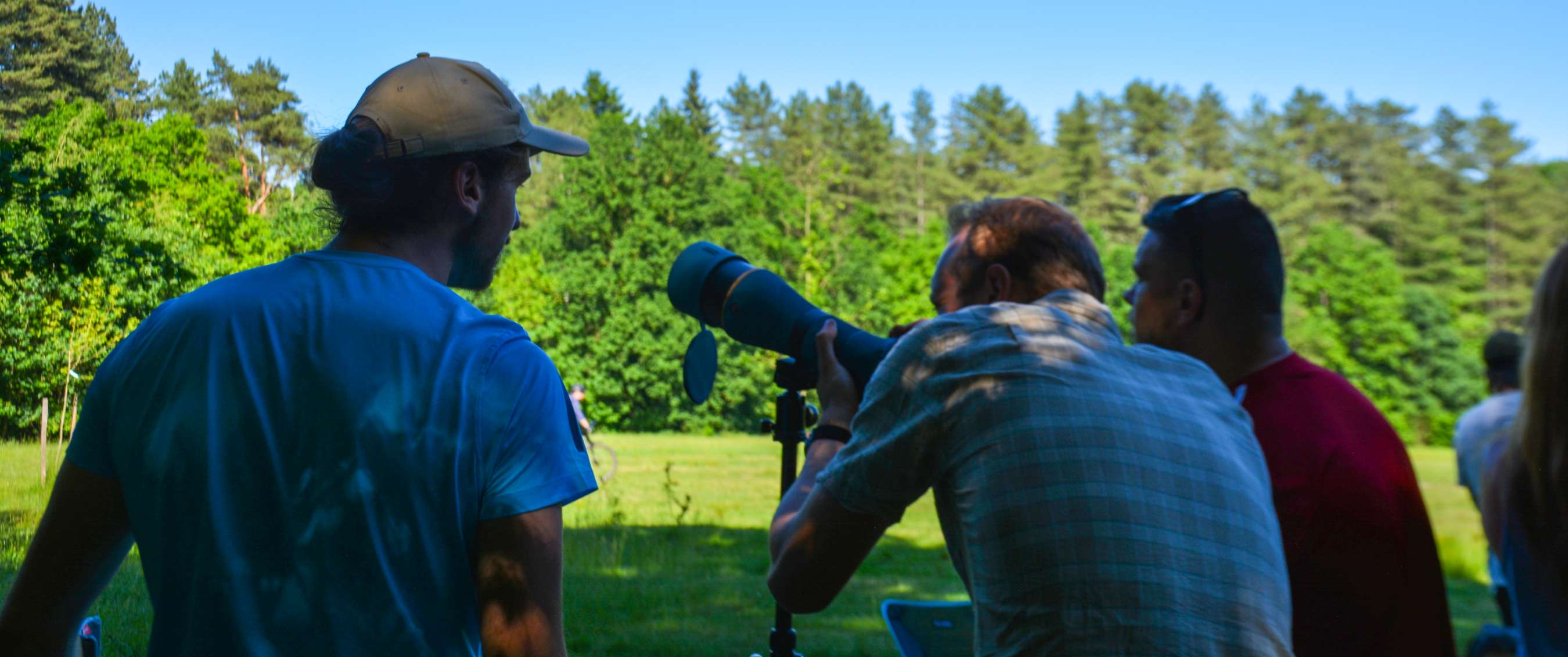 Groep observeert een groen landschap met een telescoop onder blauwe lucht.