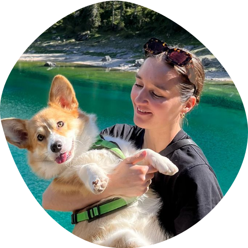 Une femme tient un corgi dans ses bras près d’un lac de montagne d’une eau bleu clair et entouré de forêts de pins. 