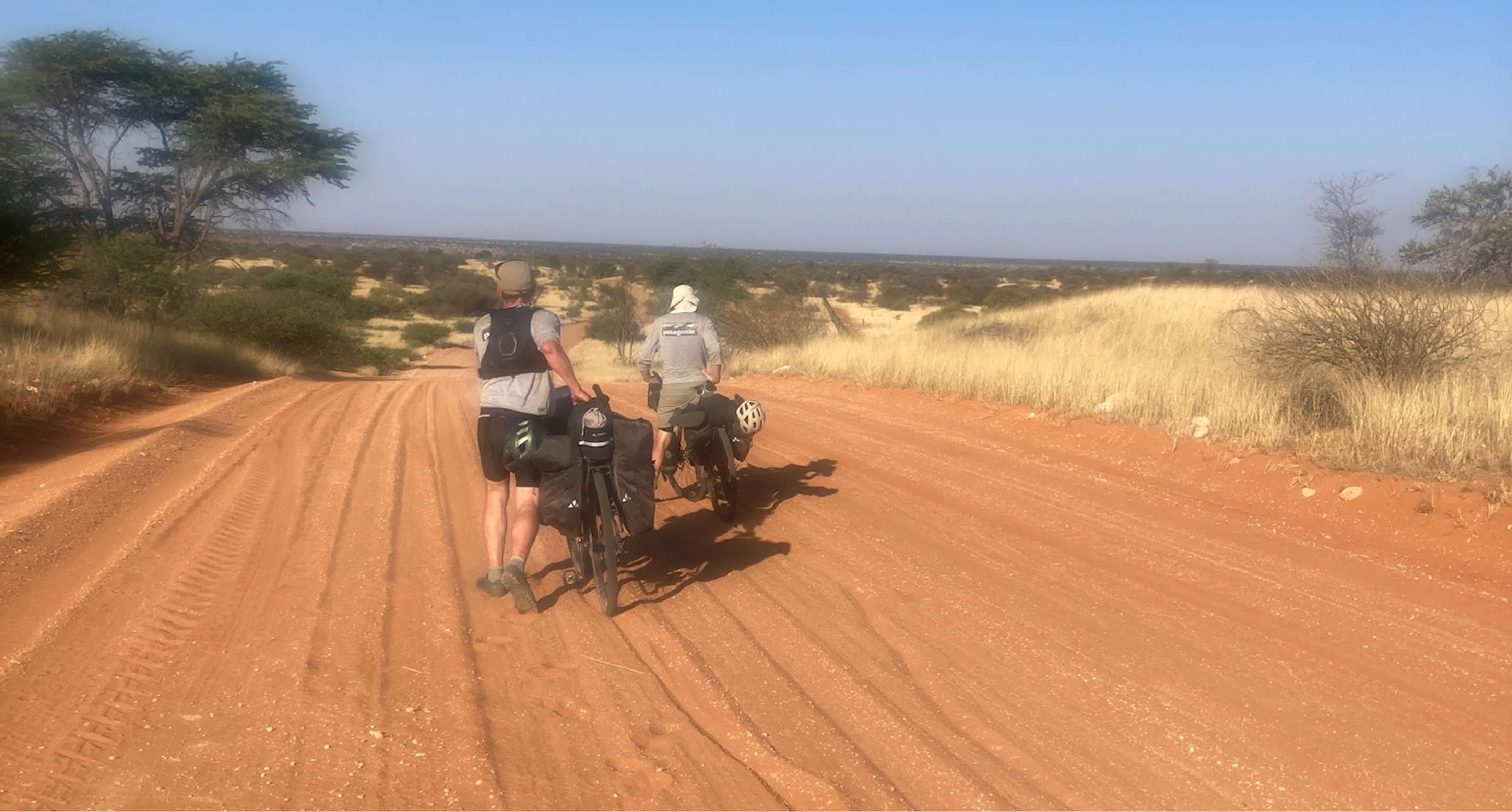 Twee fietsers duwen bepakte fietsen over een rode zandweg in droog landschap.