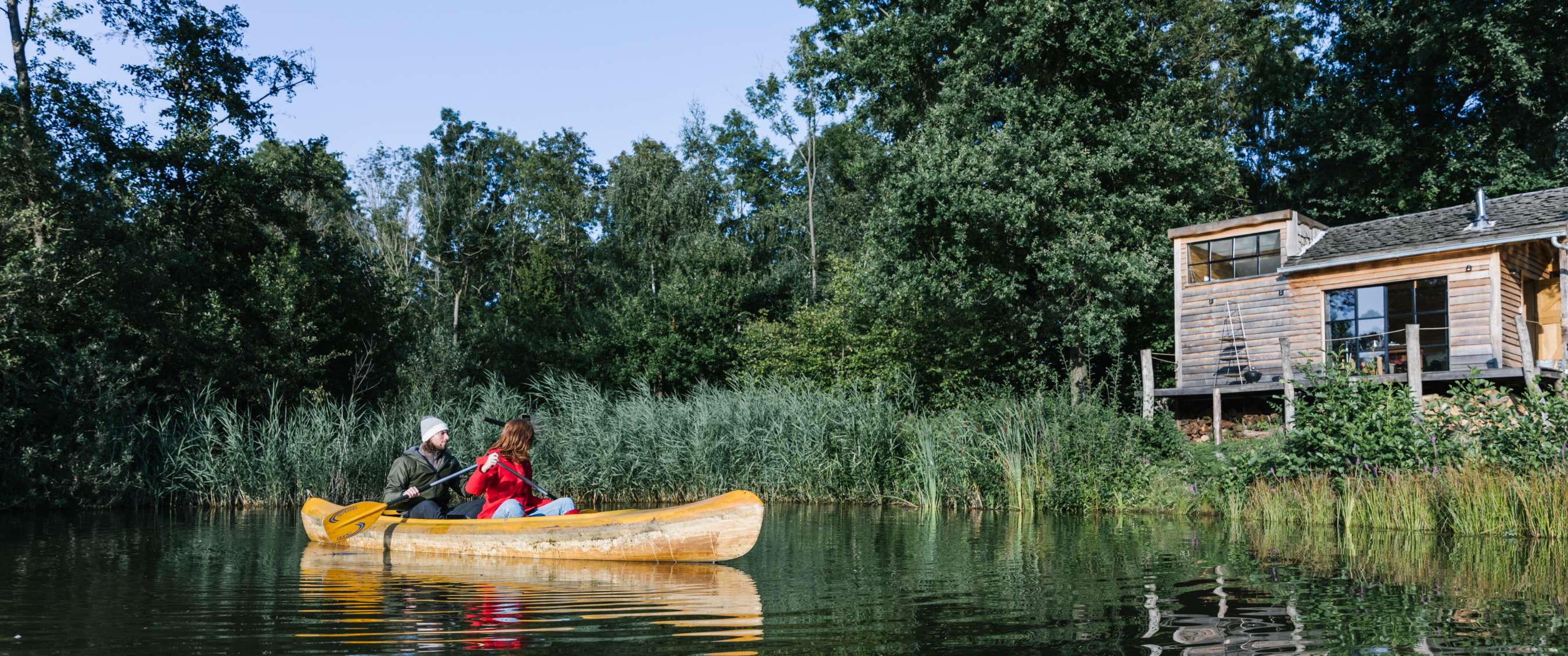 Kano met twee personen op een vijver in een groene omgeving, met een huisje zichtbaar aan de waterkant.