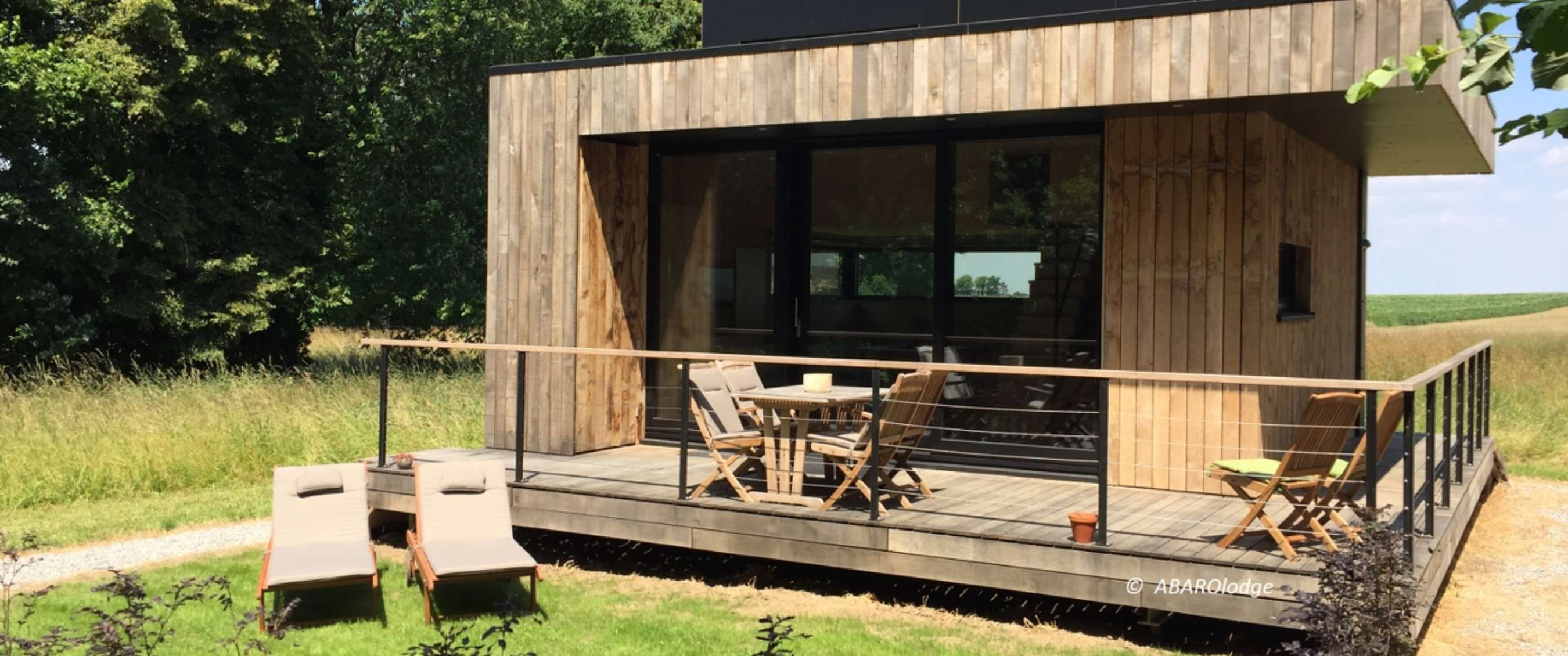 Modern houten vakantiehuis met verhoogd terras, stoelen en ligbedden in een groene omgeving.