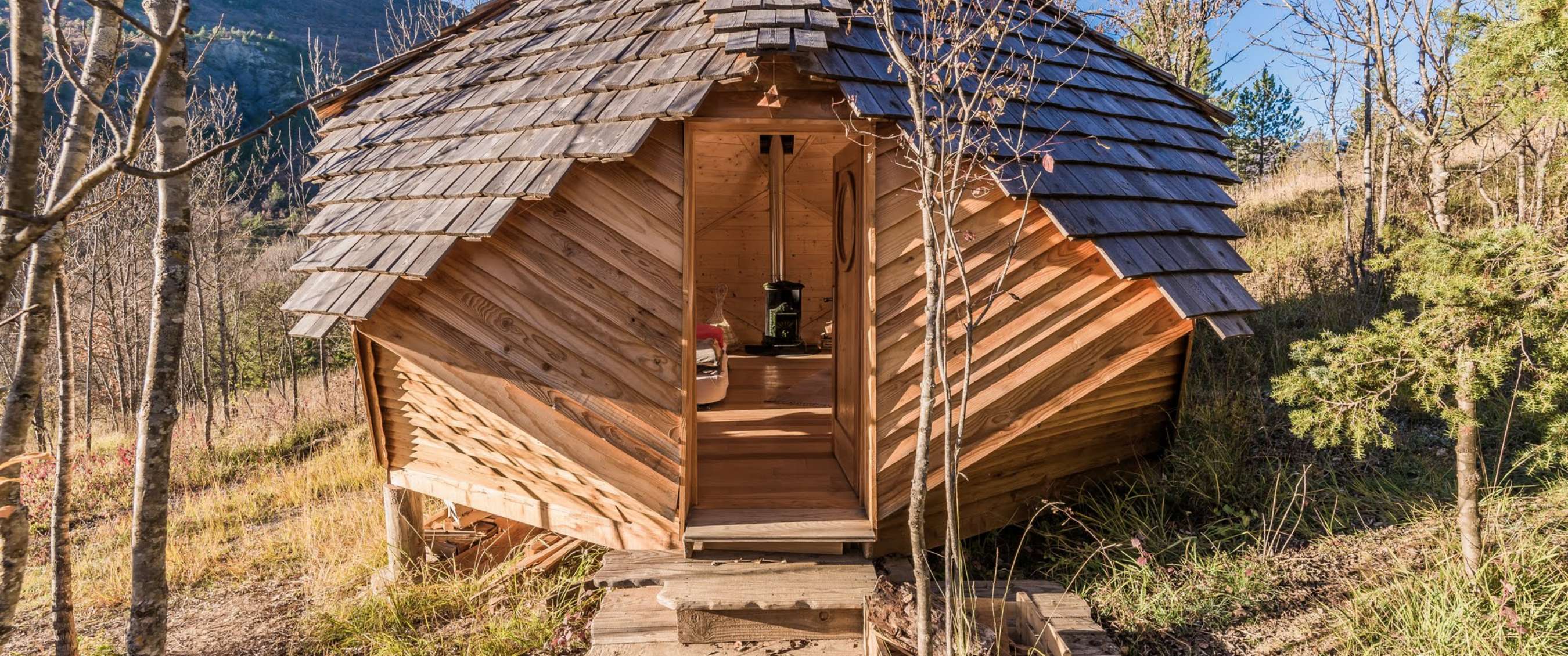 Cirkelvormig houten huisje met shingled dak in een natuurlijke bergachtige omgeving.