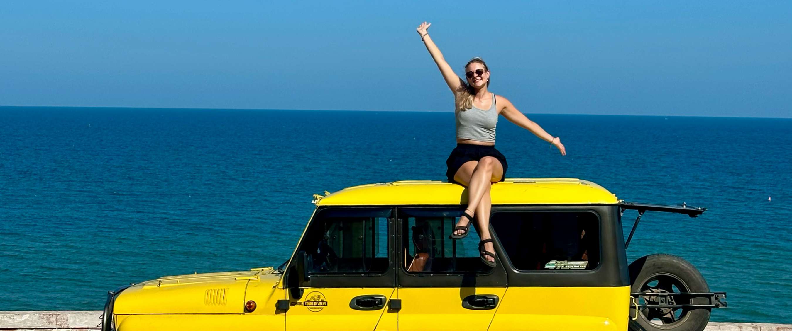 Personne assise sur une jeep jaune en saluant, avec la mer bleue en arrière-plan