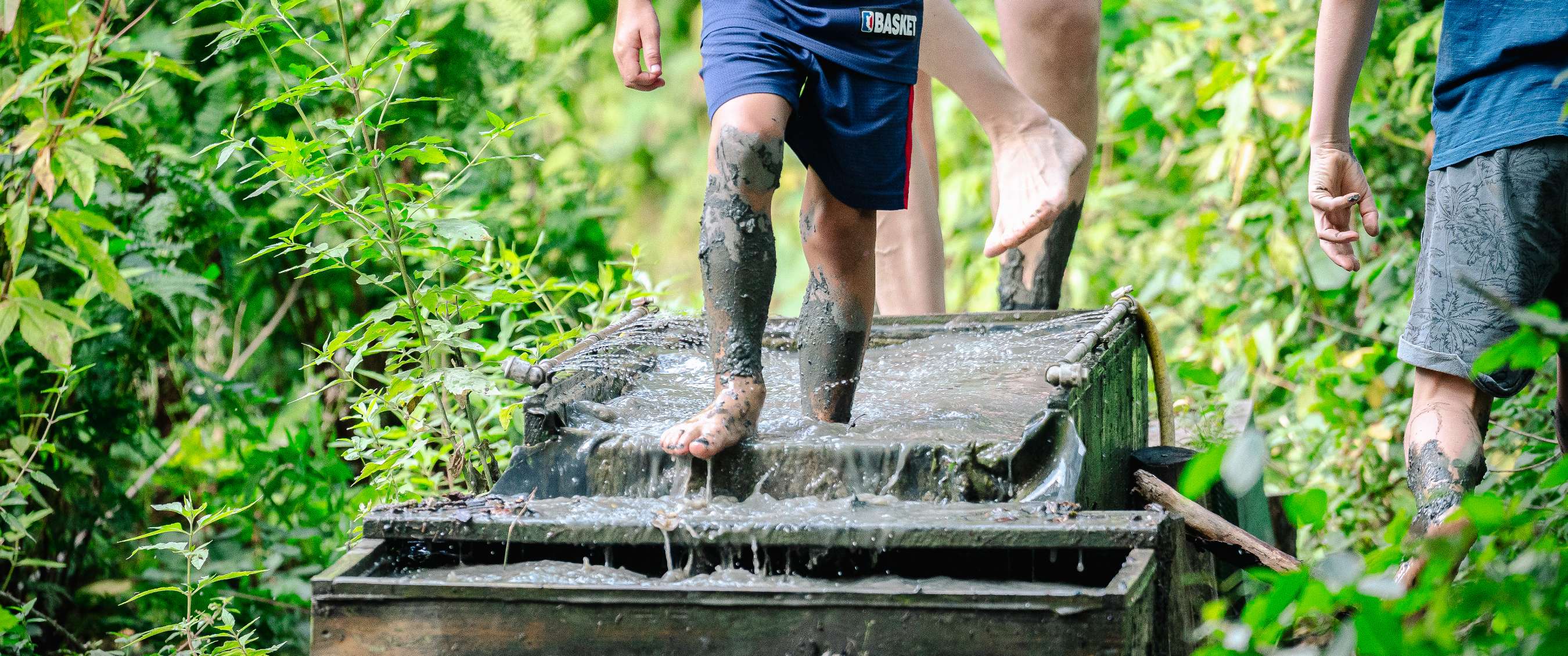 Kind loopt blootsvoets door een modderige waterbak in een groene bosomgeving.