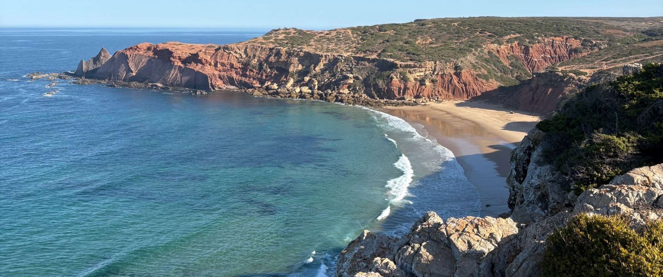 Vue sur une côte rocheuse, une plage de sable et l’océan bleu sous un ciel clair.