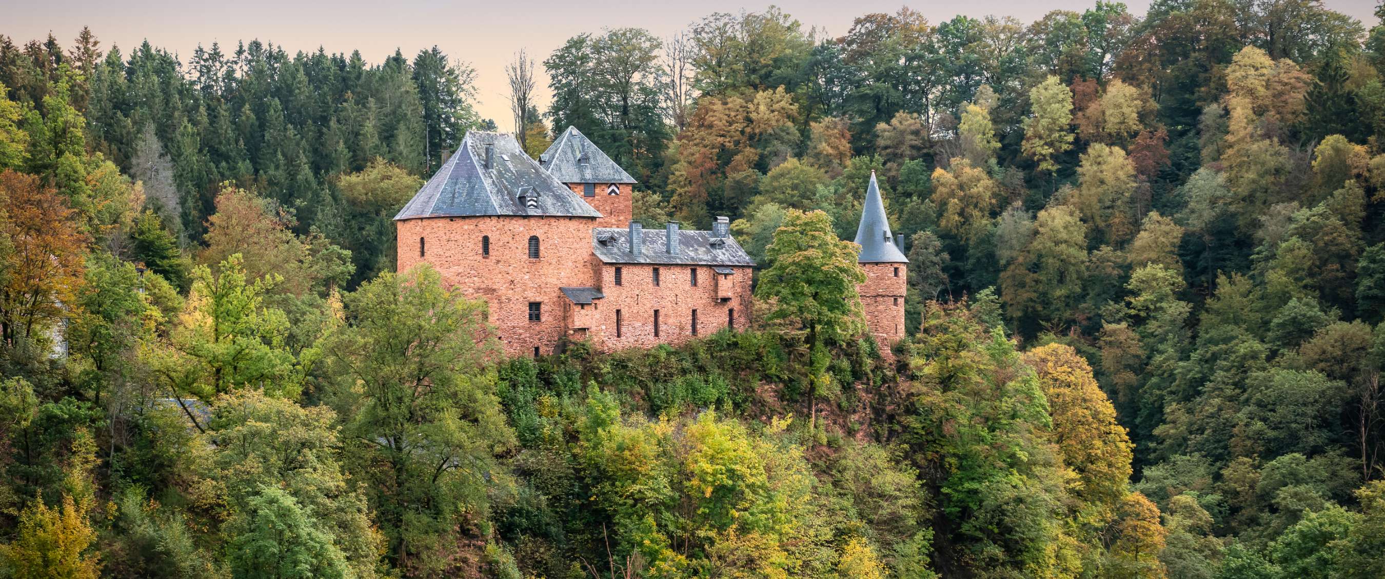 Château en pierre rouge avec tours sur une colline boisée parmi des arbres aux couleurs d’automne.