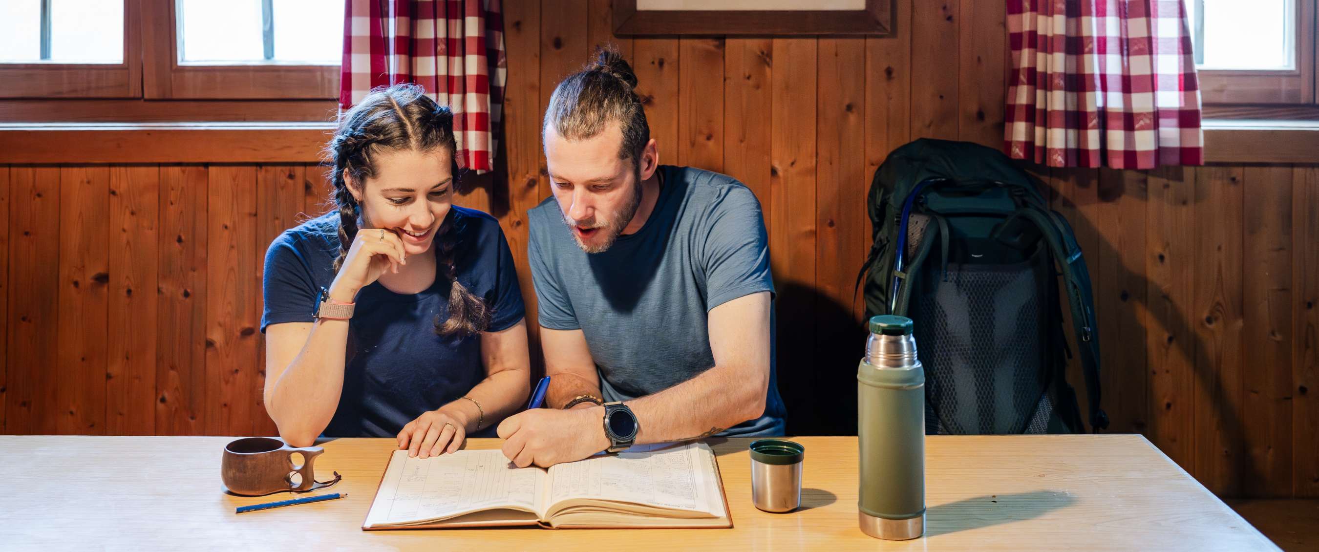 Un homme et une femme consultent une carte dans un refuge, avec un thermos et un sac à dos.
