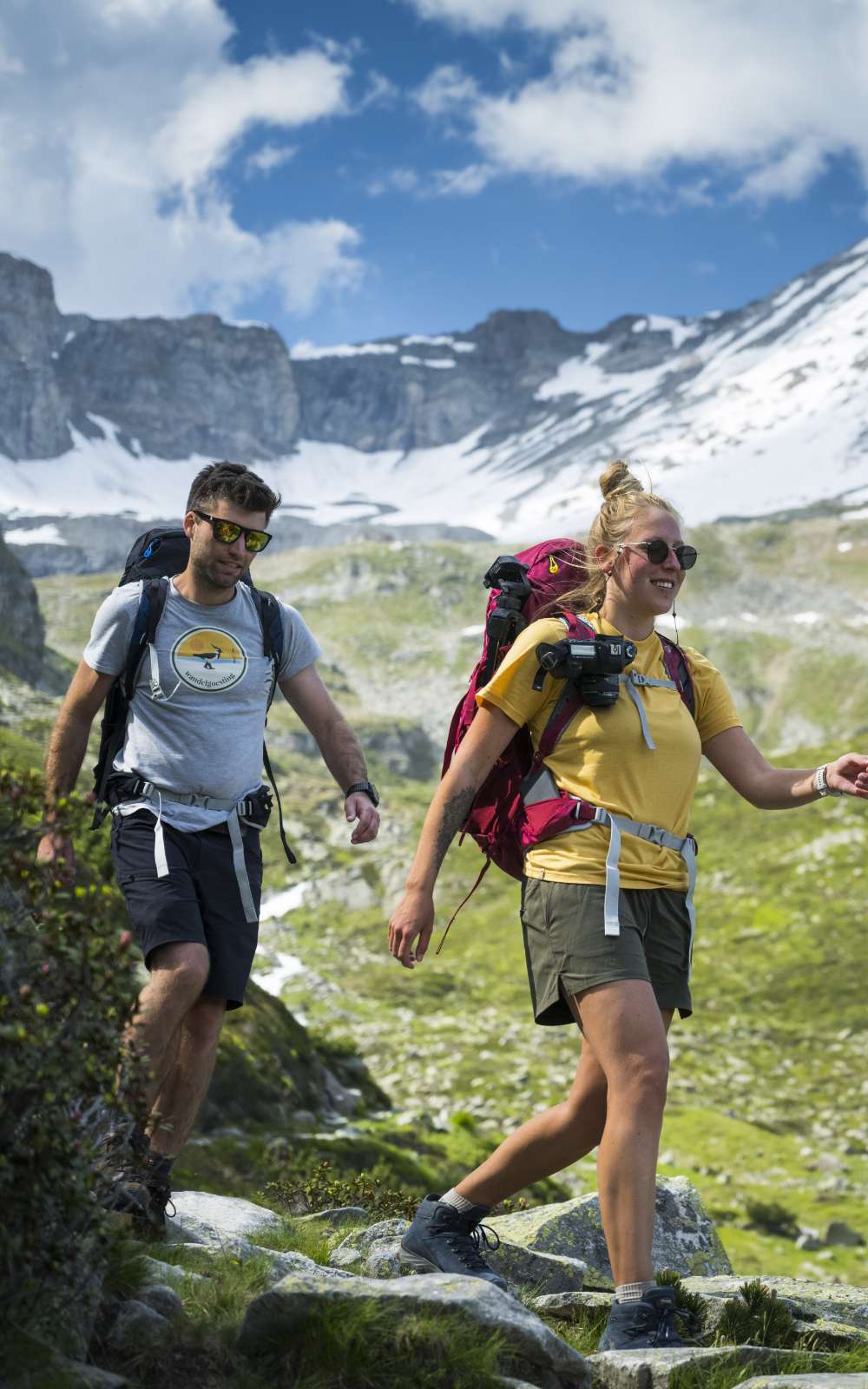 Twee wandelaars met rugzak dalen af over rotsachtig bergpad met sneeuw op de achtergrond.