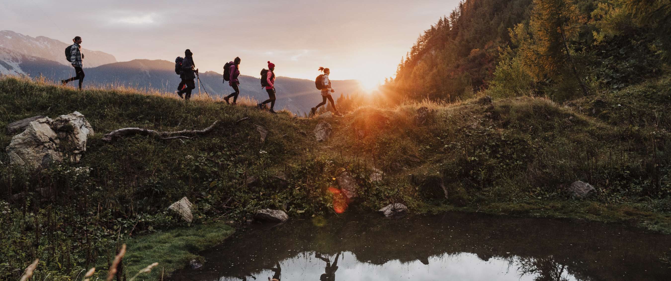 Groep wandelaars met rugzak loopt langs water bij zonsondergang in de bergen.