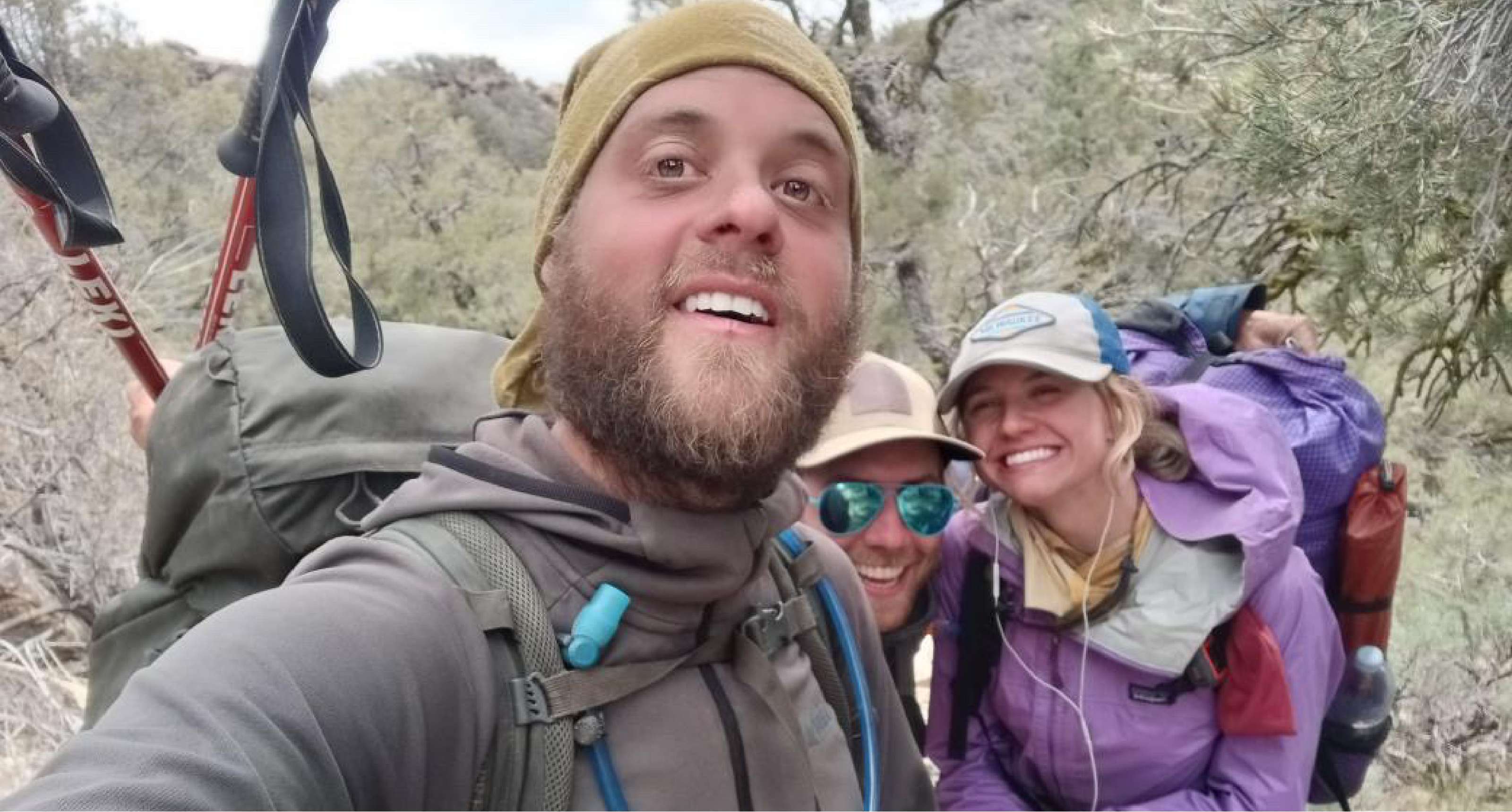 Drie wandelaars met grote rugzakken poseren samen voor een selfie in een bosrijke omgeving.
