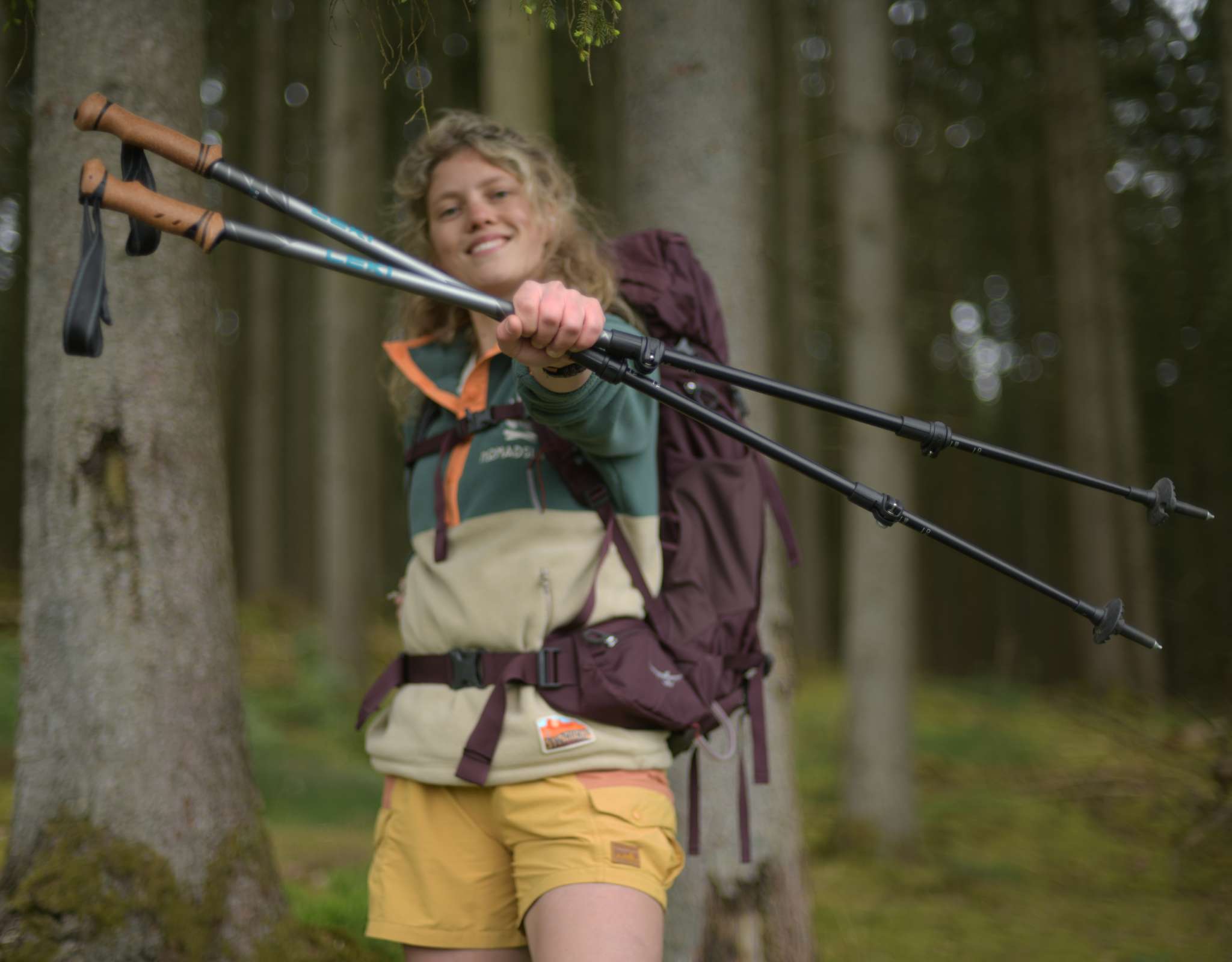Femme avec un sac à dos dans la forêt tenant deux bâtons de marche en souriant.