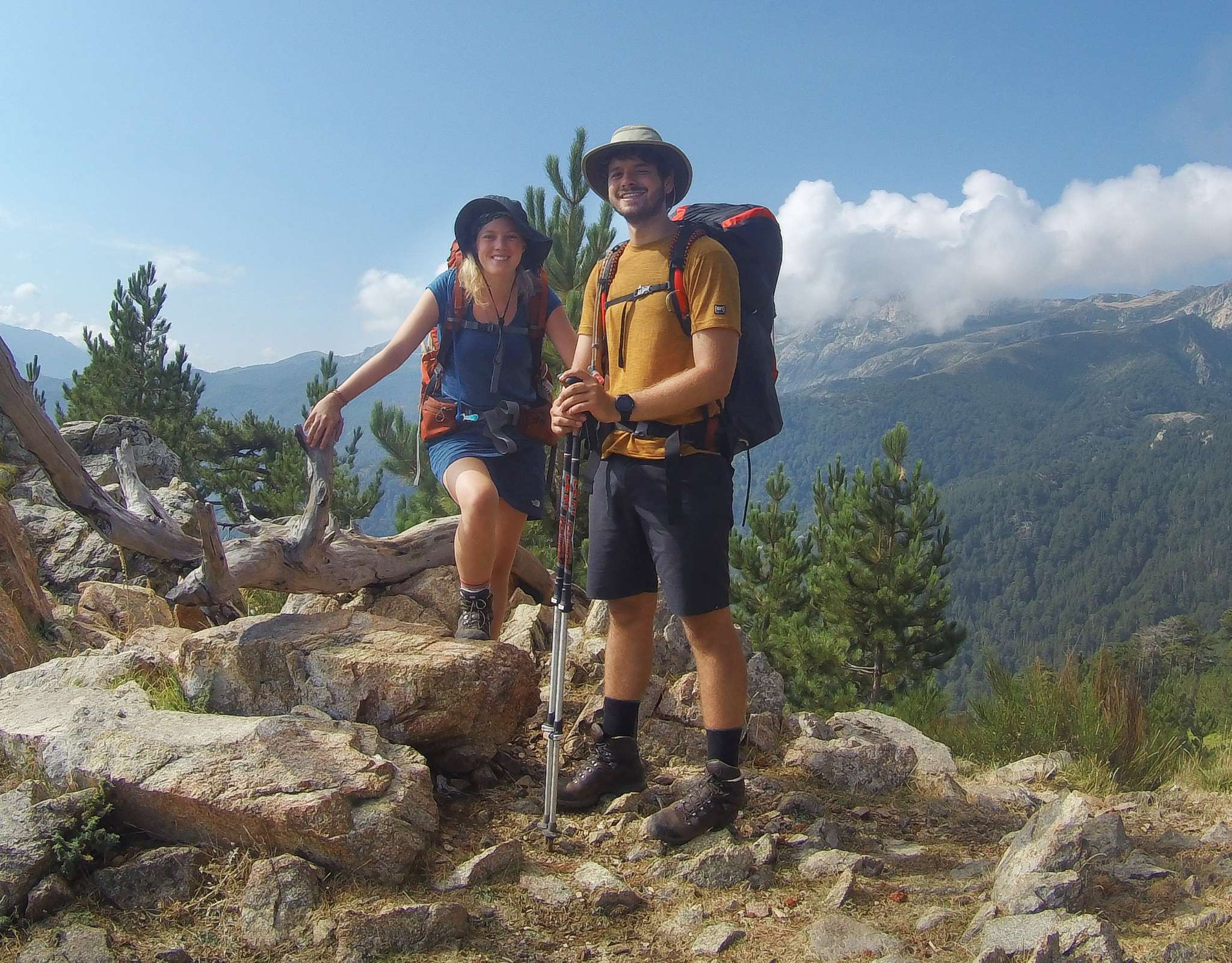 Deux randonneurs avec des sacs à dos et des bâtons de marche posent en souriant sur un sentier rocheux surplombant des montagnes verdoyantes.