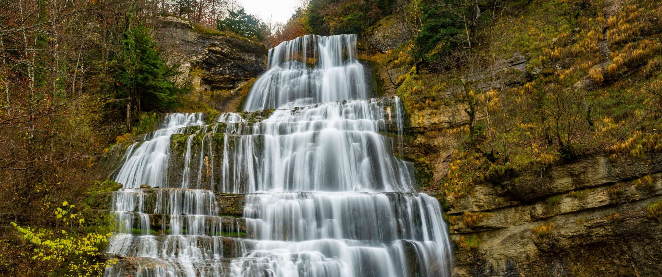 Large cascade coulant en plusieurs niveaux sur des rochers entourés d’arbres automnaux.