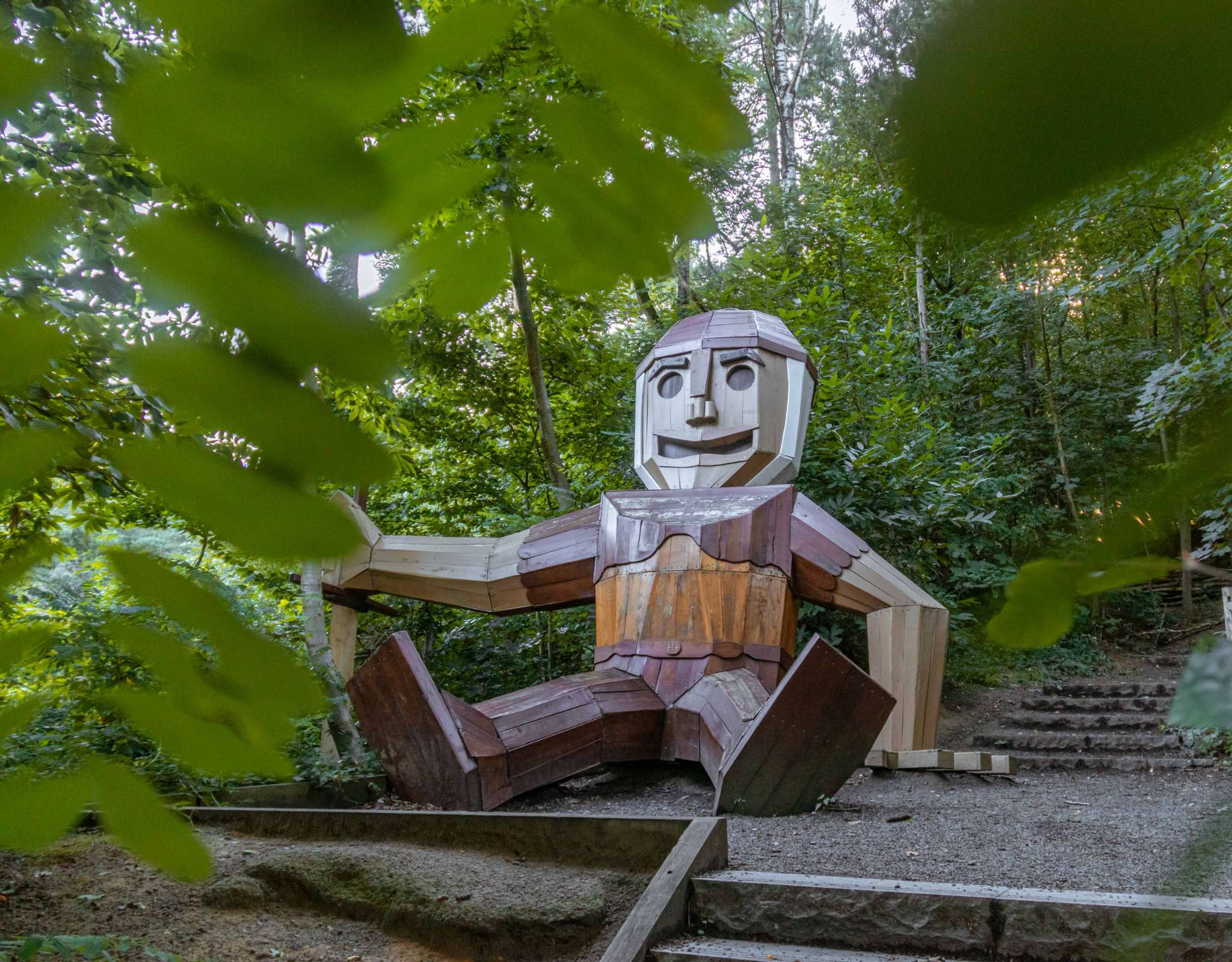 Houten sculptuur van een zittend figuur langs een bospad, omgeven door bomen