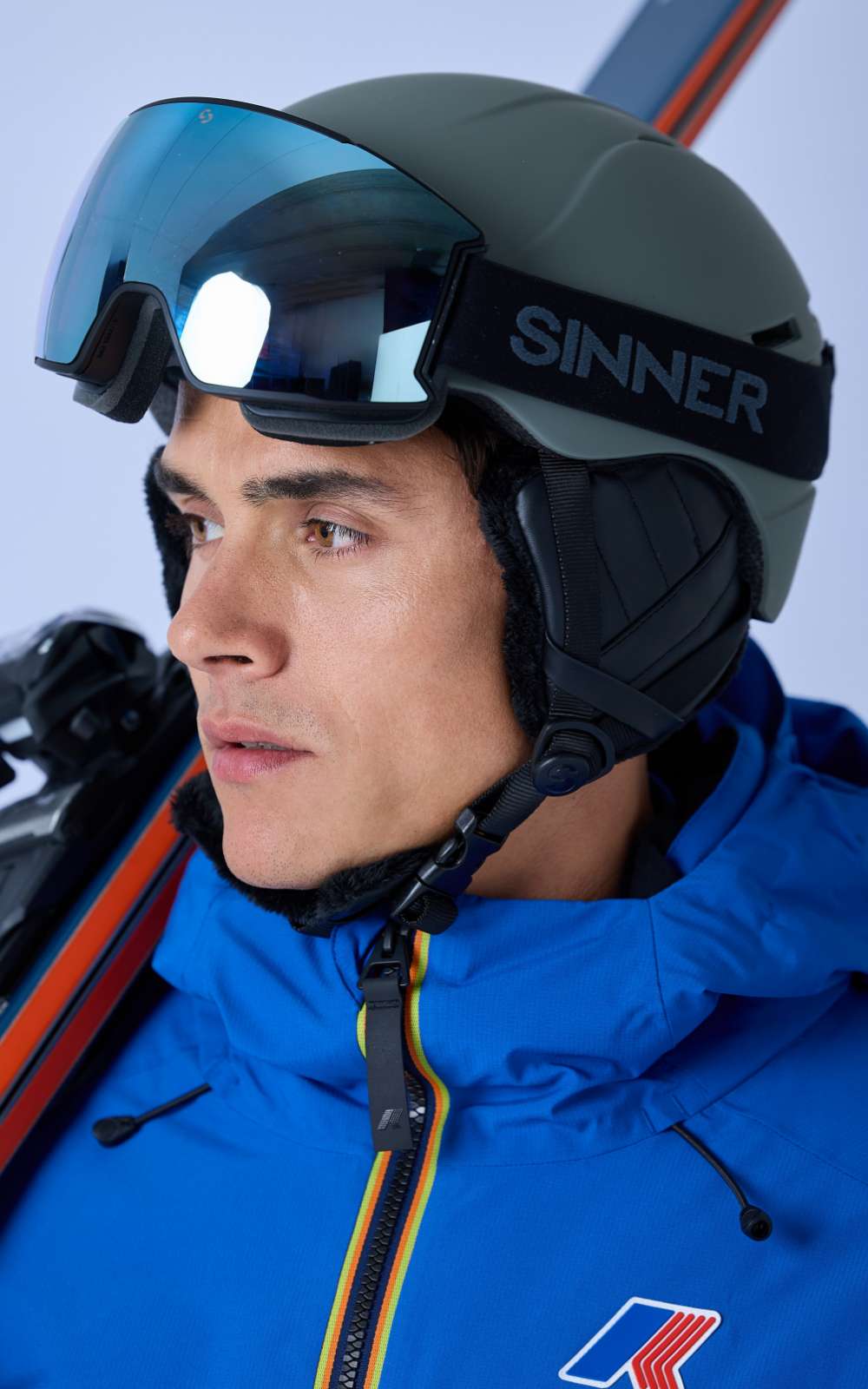 Skieur avec casque et grand masque regardant sur le côté, avec des skis visibles derrière l’épaule.