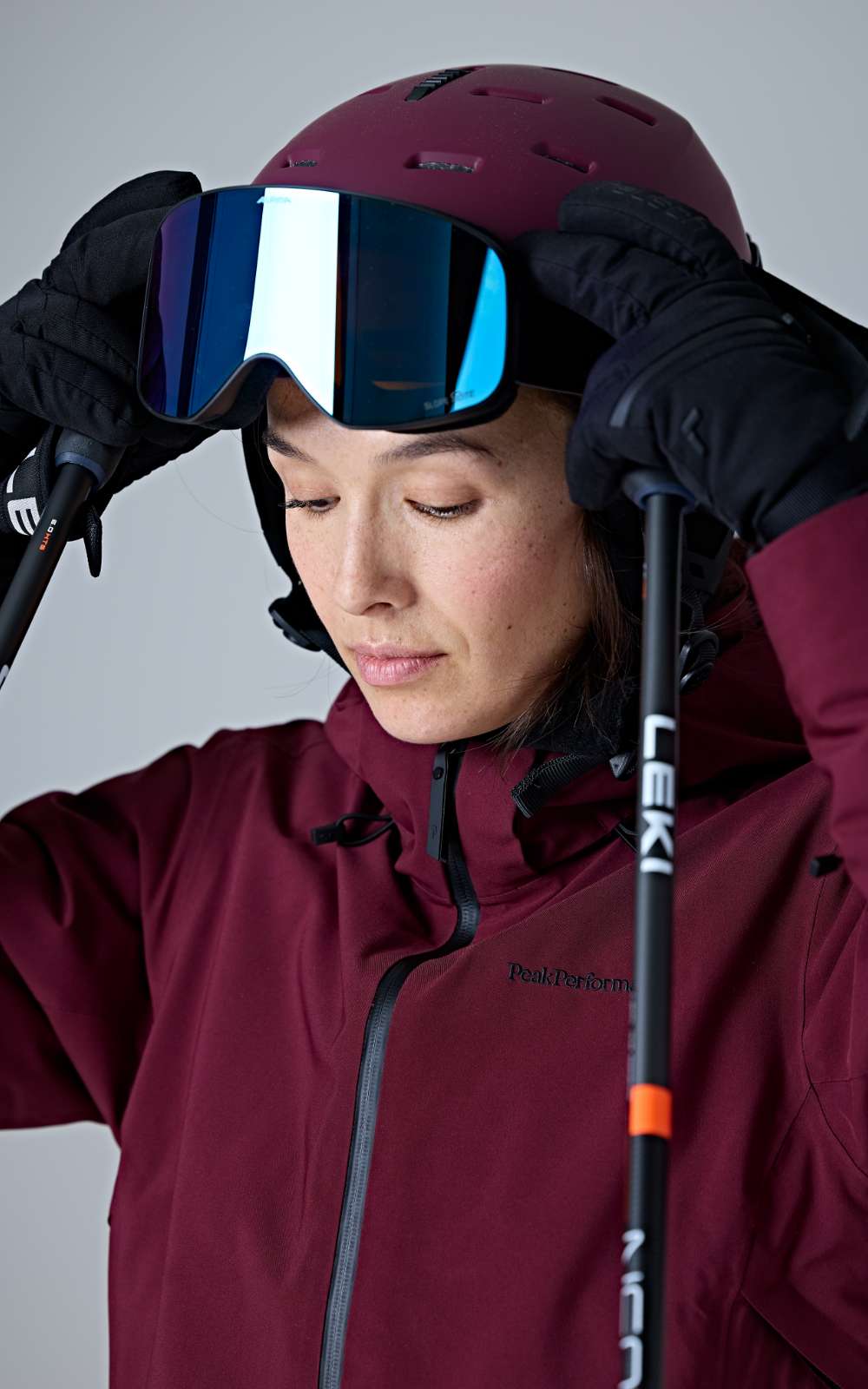 Personne en tenue de ski portant un casque bordeaux et un masque réfléchissant sur fond gris.