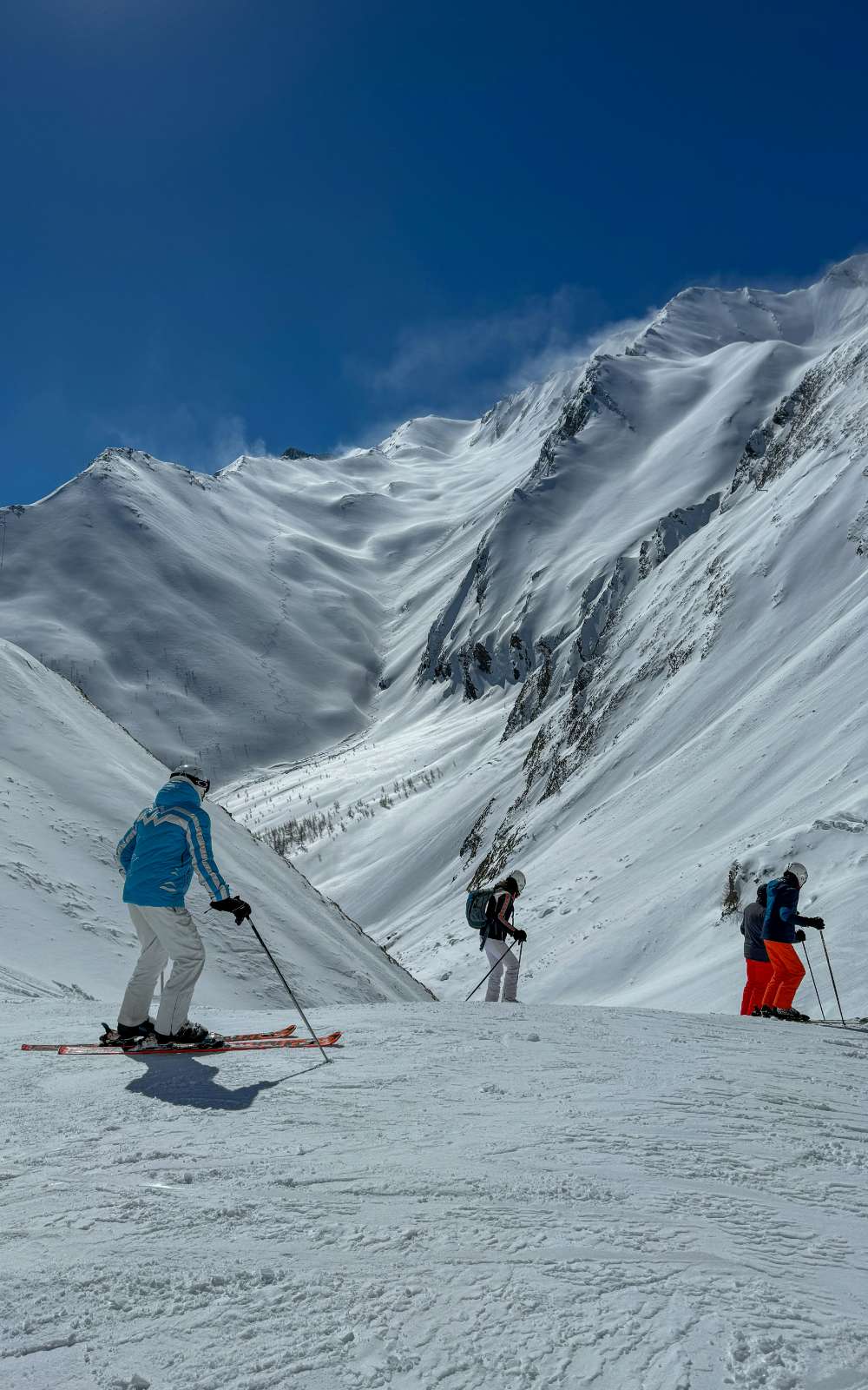Trois skieurs se tiennent sur une piste avec des montagnes enneigées escarpées à l’arrière-plan.