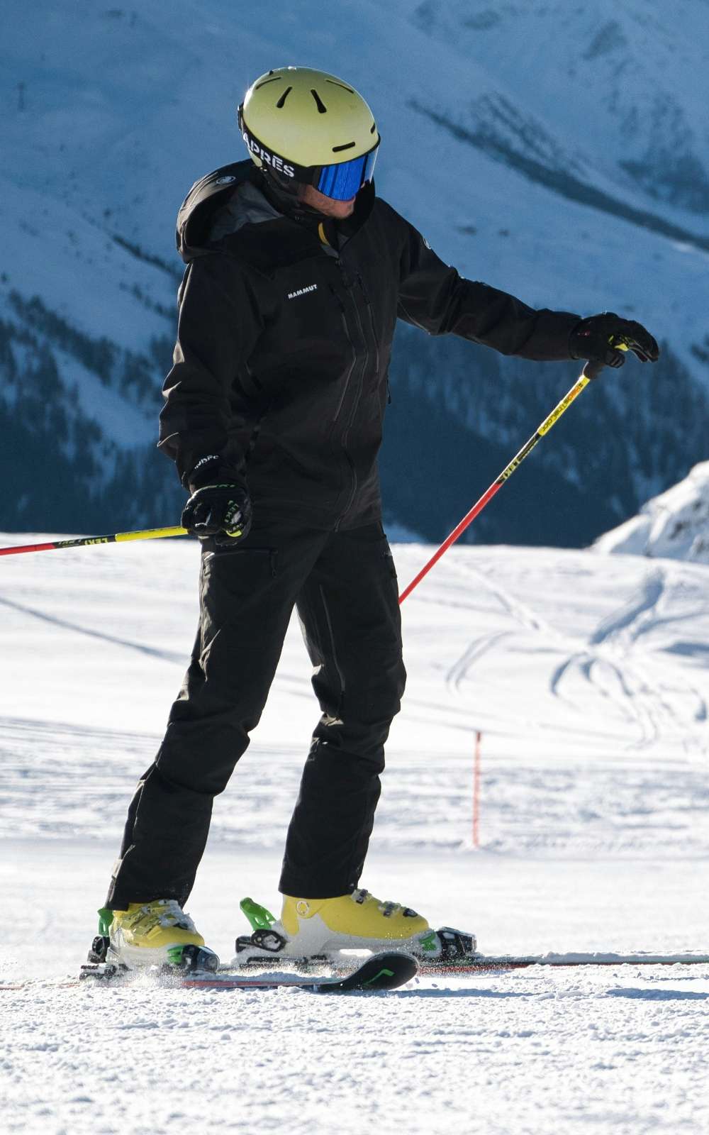 Skieur en tenue noire avec casque jaune et masque de ski tenant des bâtons sur une piste enneigée.