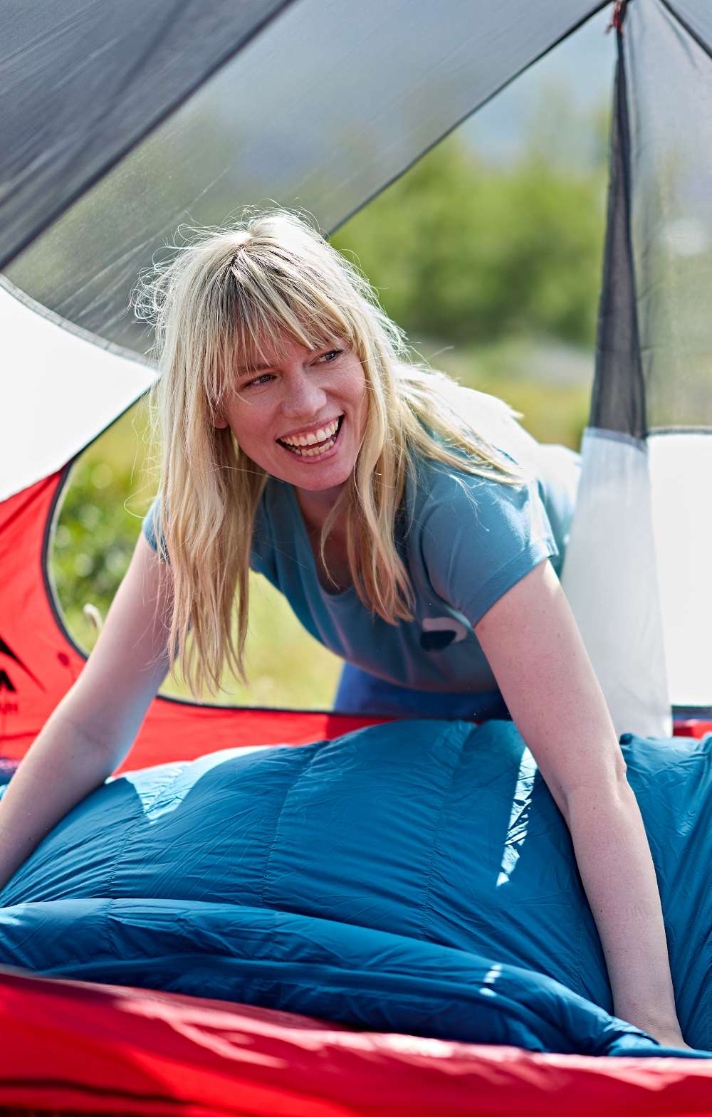 Vrouw zit glimlachend in een blauwe slaapzak in een rode tent