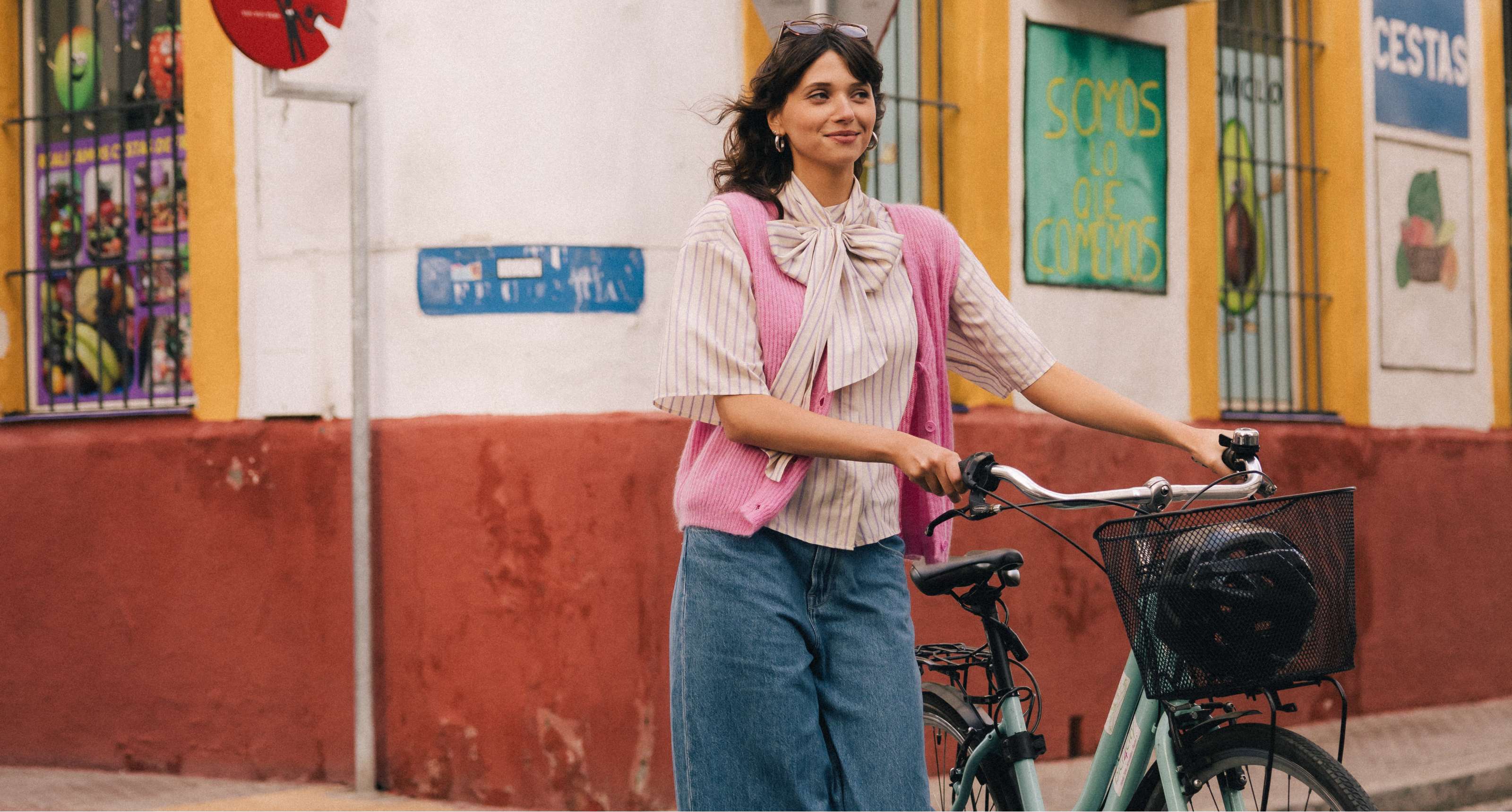 Vrouw in wijde jeans en roze gilet loopt met een fiets langs een kleurrijke gevel.
